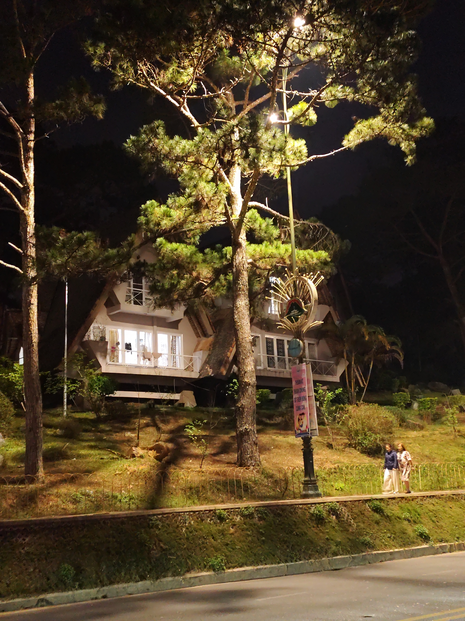 At night in Da Lat, Vietnam, a two-story building with white walls and brown trim is visible behind trees. The building has an A-frame structure, with lighted windows on both floors. A balcony is visible on the lower level. A sign on a pole stands in front of the building. Two people stand on the sidewalk. The sky is dark.