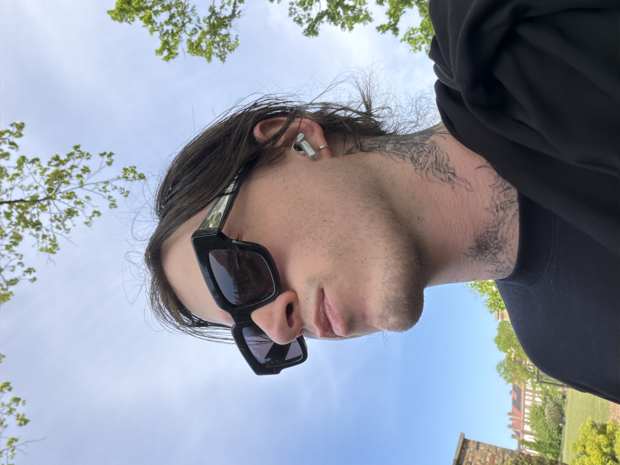The image, oriented with the sky at the top, captures a close-up, upward-angled view of a person outdoors. The subject, seen in profile, is a person with dark, medium-length hair, wearing square-framed black sunglasses and a silver earbud in their right ear. Facial stubble is visible on their chin and jawline. Elaborate black tattoos extend from below their right ear down their neck and onto their shoulder, partially covered by a plain black top, possibly a t-shirt or hoodie.
The person appears to be looking slightly downwards or away from the camera, giving a casual and contemplative impression. The sky above is clear and bright blue, suggesting a sunny day. Green tree branches with fresh leaves frame the top and sides of the frame, indicating a natural, outdoor setting, likely a park or garden. In the distance, on the right, a multi-story building with reddish brickwork and numerous windows can be seen, hinting at an urban or suburban environment.
Visible text on the arm of the sunglasses appears to be "BALENCIA", possibly indicating the brand. Given the location context, this scene is set in London, United Kingdom.