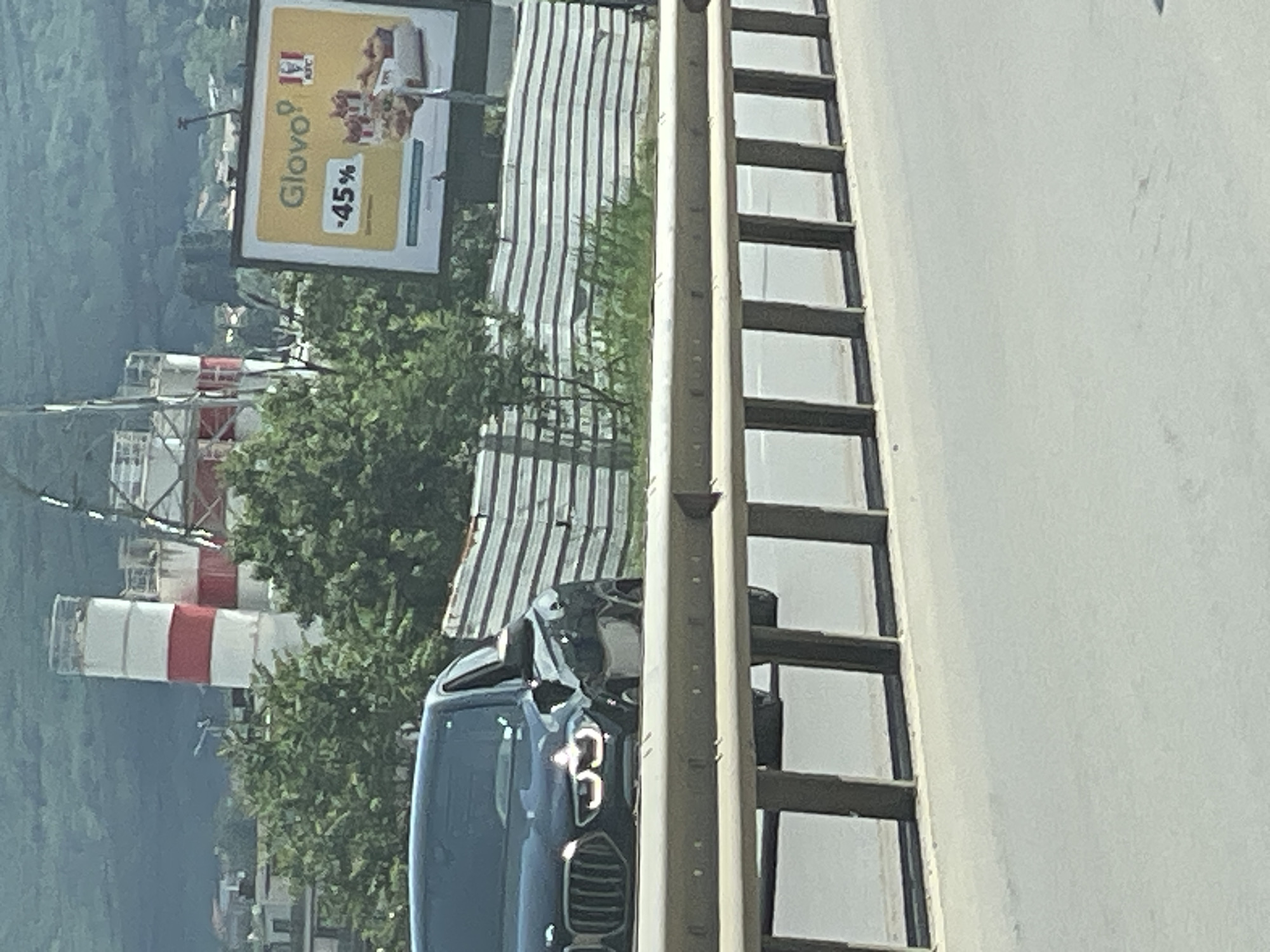 The image shows a black BMW driving on a highway. In the background, there is a billboard advertising Glovo and KFC with a "45%" discount. There are also industrial silos in the background. The scene appears to be in Sofia, Bulgaria.