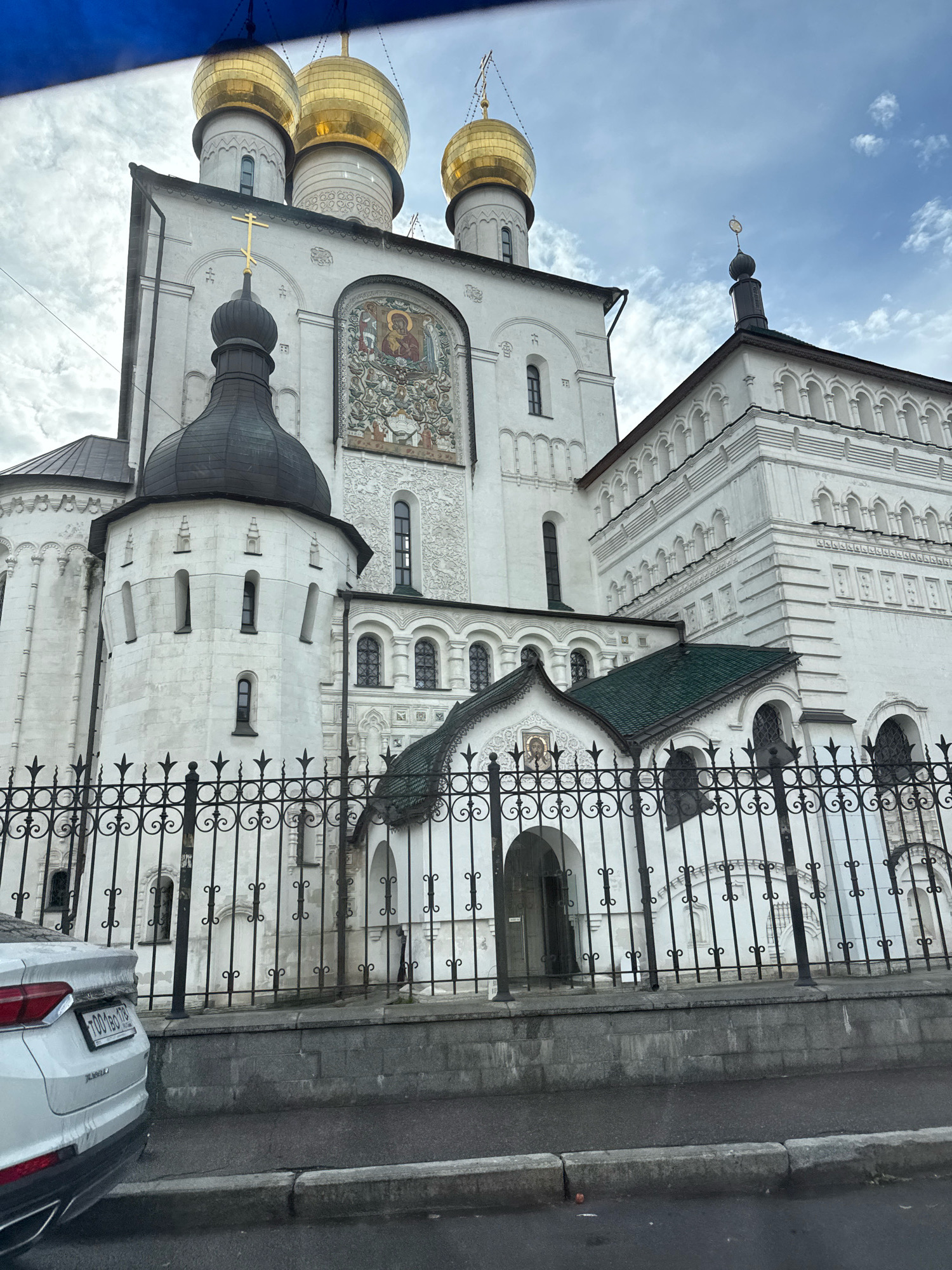 The image depicts the Church of the Intercession of the Virgin Mary in Fili, Moscow, Russia. The church is a white stone building with golden domes and a black onion dome. It is surrounded by a black wrought-iron fence. In the foreground, the rear of a white car is visible. The sky is cloudy.