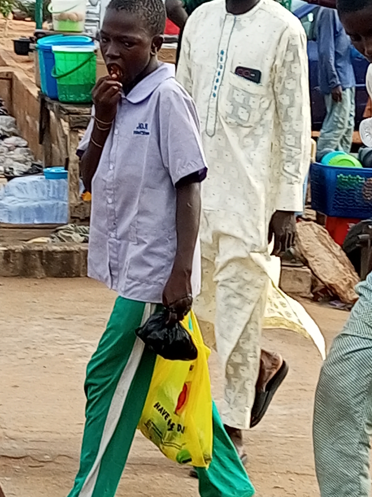 The image shows a young boy in the foreground, wearing a light purple collared shirt with blue trim and a school emblem, and green pants with a white stripe. He is eating something from his hand. He is carrying a black plastic bag in his left hand and a bright yellow shopping bag in his right, which has some text on it. Behind him, a man in a light-colored, patterned traditional tunic walks away from the camera. The setting appears to be an outdoor market or roadside, with various containers and objects visible in the background, including blue and green buckets and a blue plastic crate. The ground is sandy or dusty. The overall impression is of a busy, everyday scene, possibly in the afternoon given the lighting. The boy's expression is neutral, focused on his food. The text on the yellow bag is partially visible and appears to read "HAVE A DI...". The scene is likely in Gombe, Nigeria.