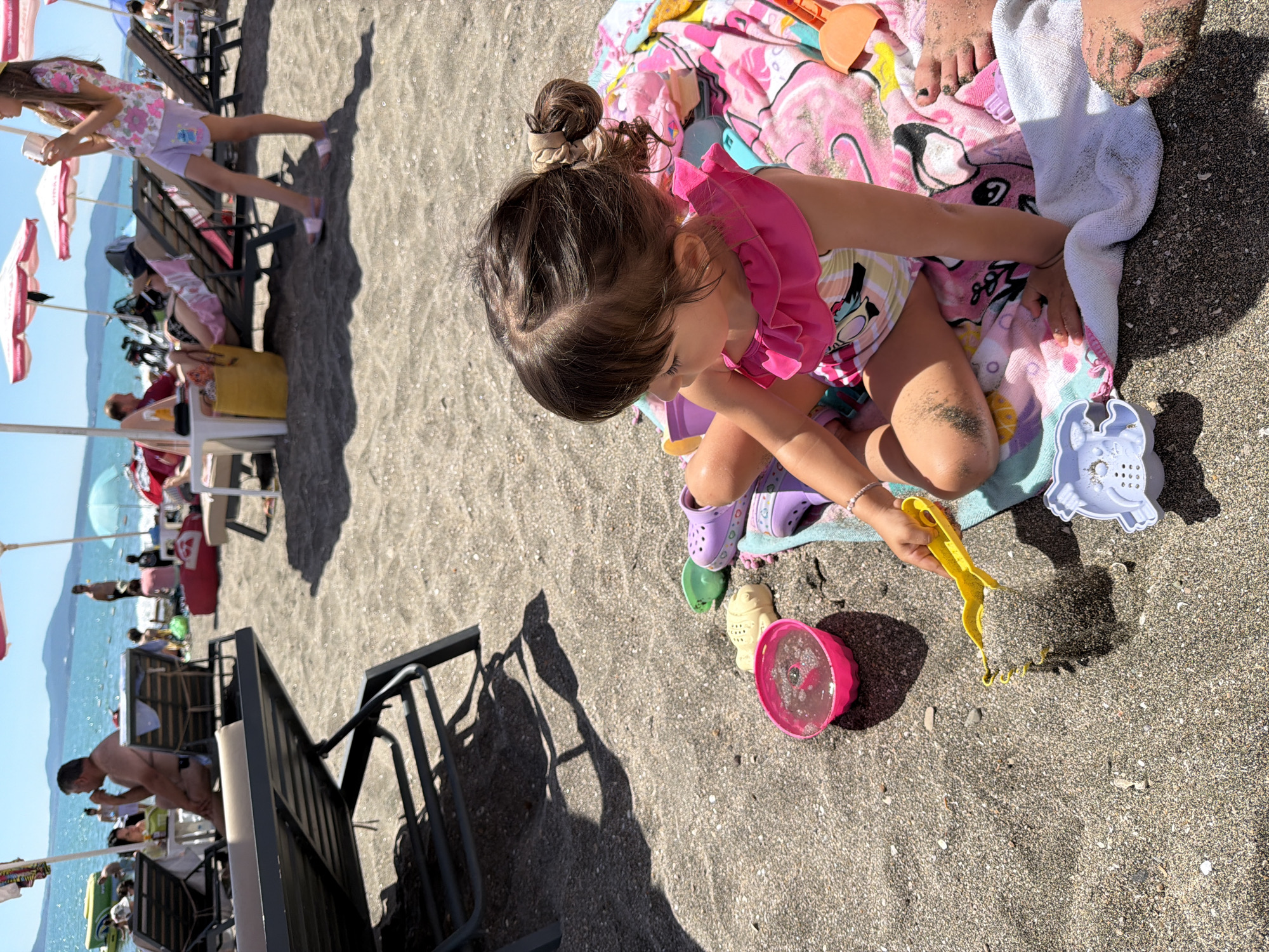 The image depicts a sunny day at a beach in Burgas, Bulgaria. The foreground is dominated by a young girl with her hair in a bun, wearing a pink swimsuit and purple Crocs. She is sitting on a colorful beach towel with a cartoon print, surrounded by sand toys. The girl is actively playing with a yellow sand rake, scooping sand into a pink bucket. Next to her are other sand molds, including a yellow fish and a white crab-shaped one. In the background, the beach is dotted with beach chairs and umbrellas, with people enjoying the seaside. The turquoise water of the sea is visible, and mountains form a distant backdrop. The sunlight creates strong shadows on the sand, indicating it is likely midday. The overall atmosphere is one of leisure and childhood fun. There is no visible text in the image.