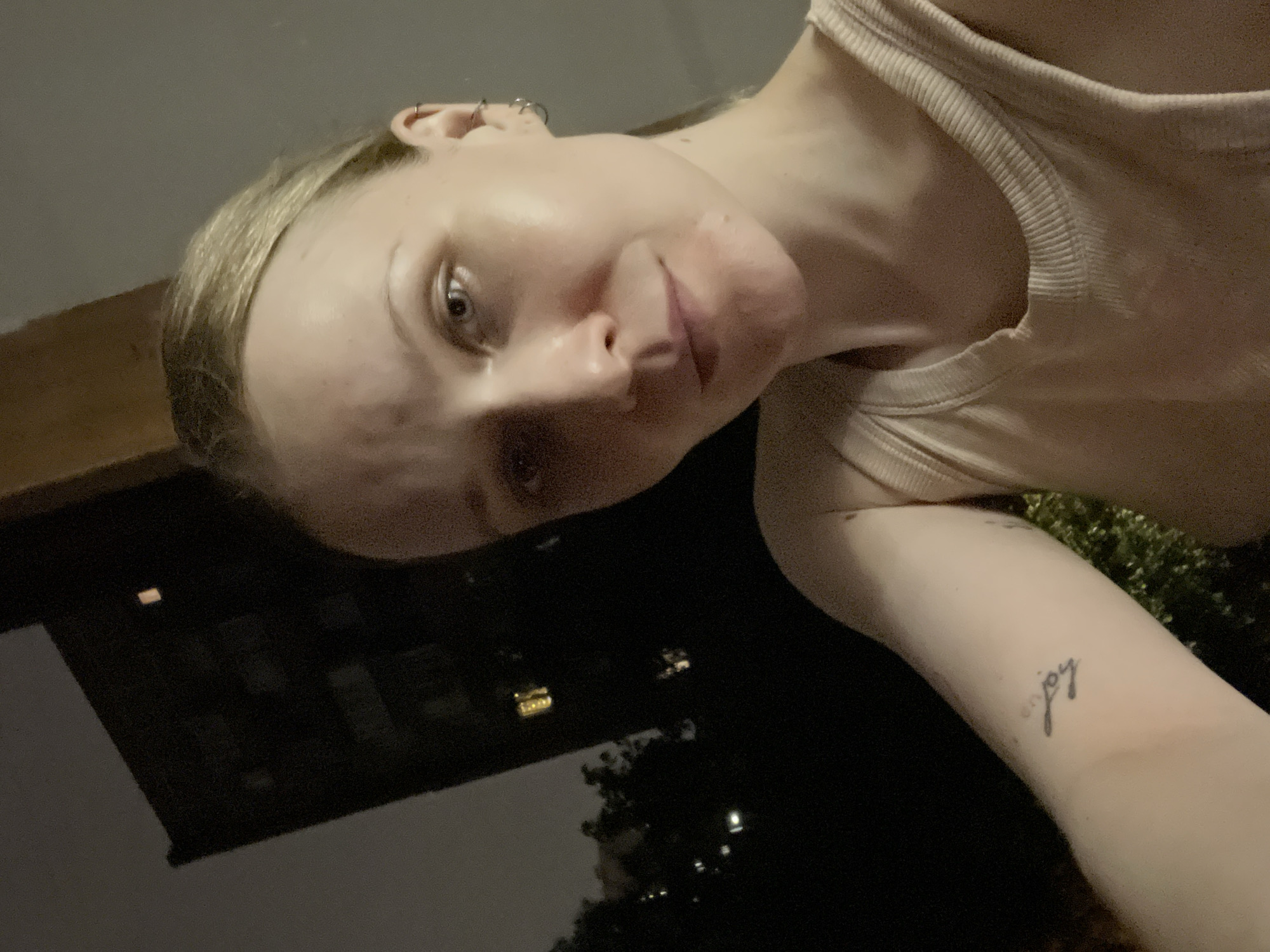 A medium shot presents a blonde woman with fair skin, wearing a light-colored tank top. She is positioned outdoors, with a building visible in the background, possibly in a city environment. The lighting suggests it might be nighttime. There is a tattoo reading "joy" on her arm. The scene lacks precise geographical indicators.
