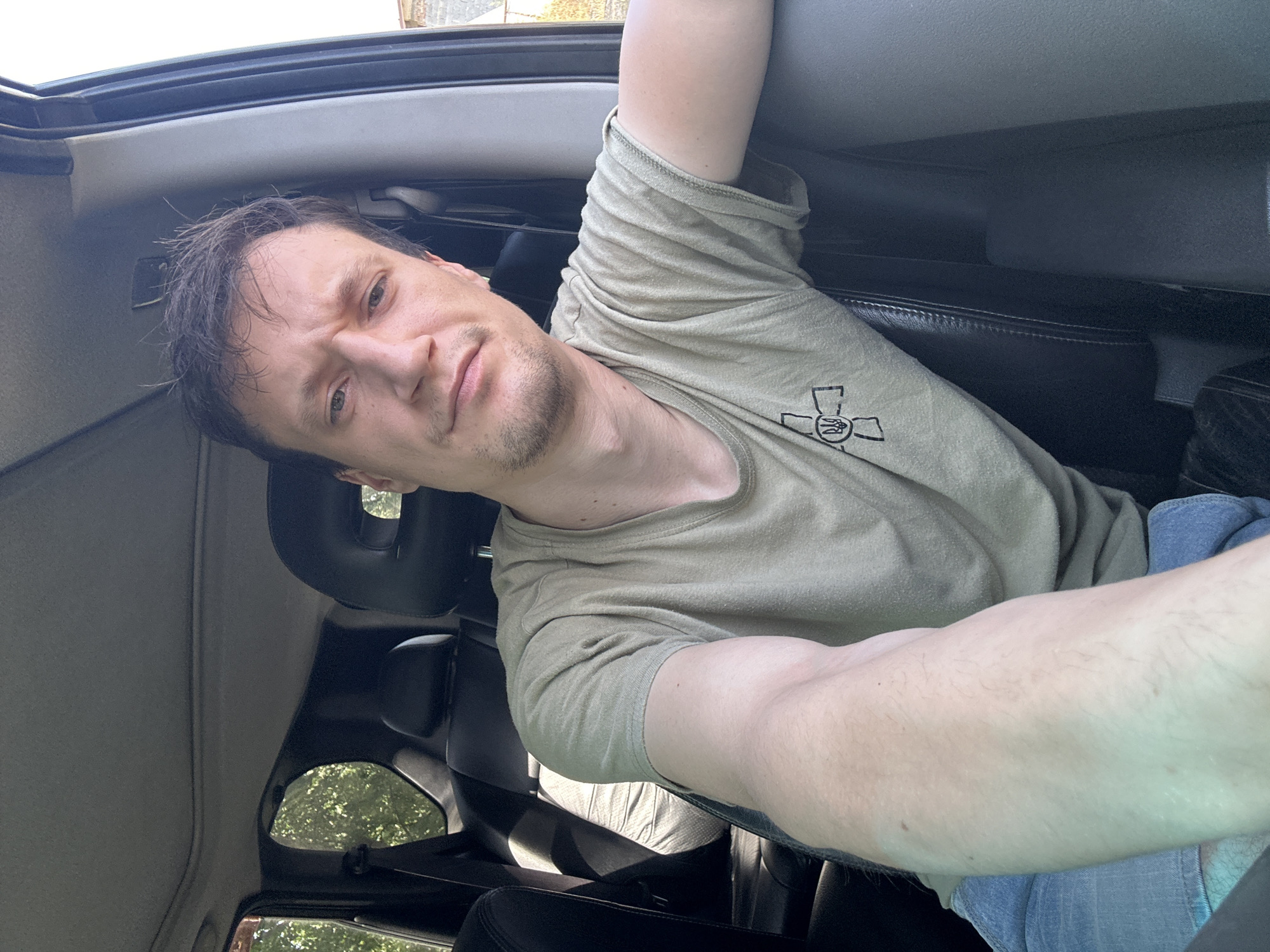 A man with short, dark hair and light skin is sitting in the driver's seat of a vehicle, looking towards the camera. He is wearing a light green t-shirt with a black symbol on the left breast and blue jeans. His left arm is extended towards the camera, taking a selfie. The interior of the car is visible, including black leather seats and a sunroof. Outside the car, trees and a building can be seen through the windows. The image appears to have been taken during the daytime.