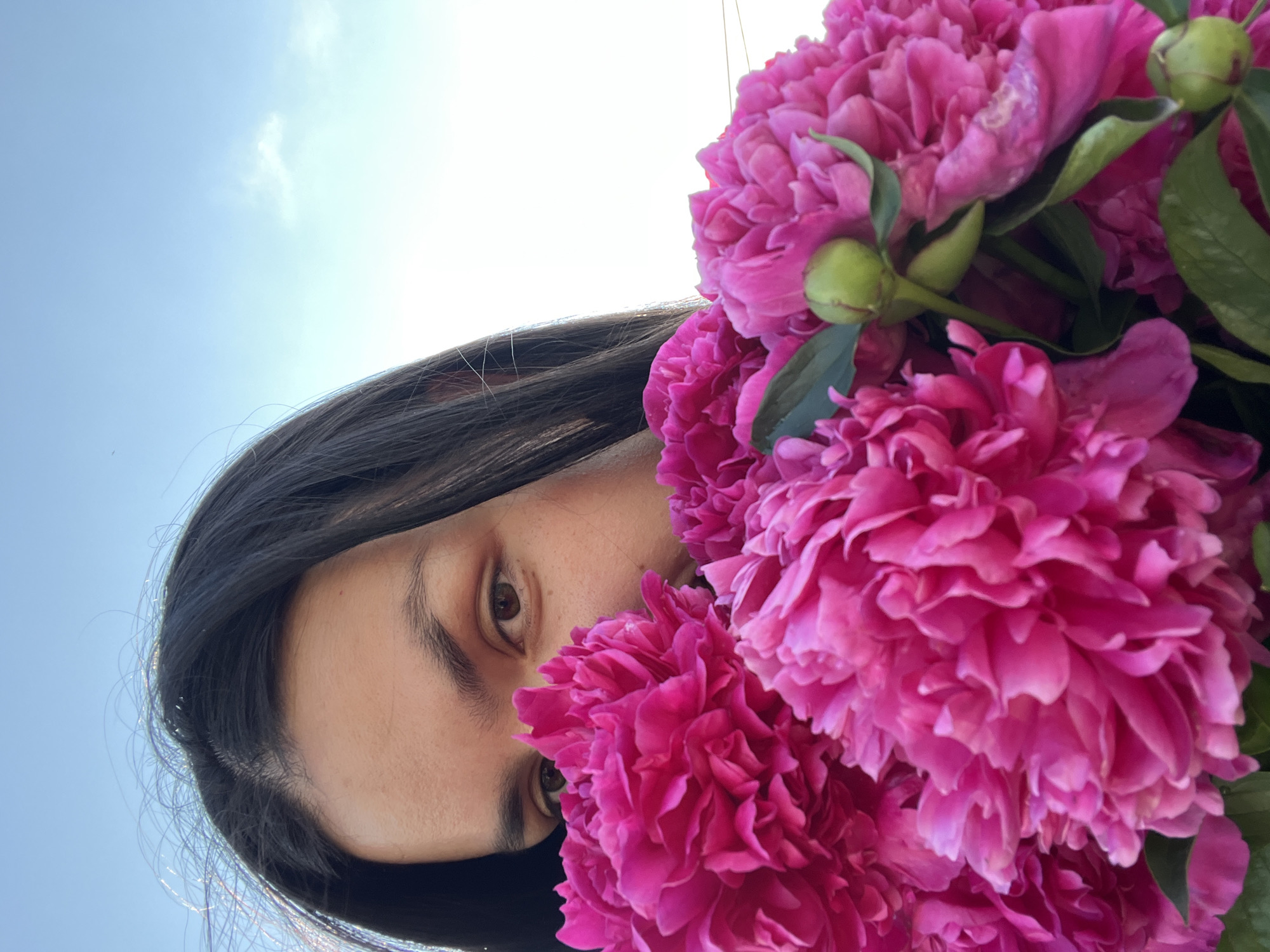 An extreme close-up, low-angle shot captures a young woman partially obscured by a vibrant bouquet of bright pink peonies, held up to her face. Her dark hair cascades down, framing a portion of her face, with her left eye and eyebrow clearly visible. The background consists of a bright, pale blue sky with a few wispy white clouds. The lighting suggests an outdoor setting during the daytime, possibly in Oktyabrsky, Russia, given the context. The focus is sharp on the flowers and the woman's visible features, creating a sense of intimacy and vibrancy.