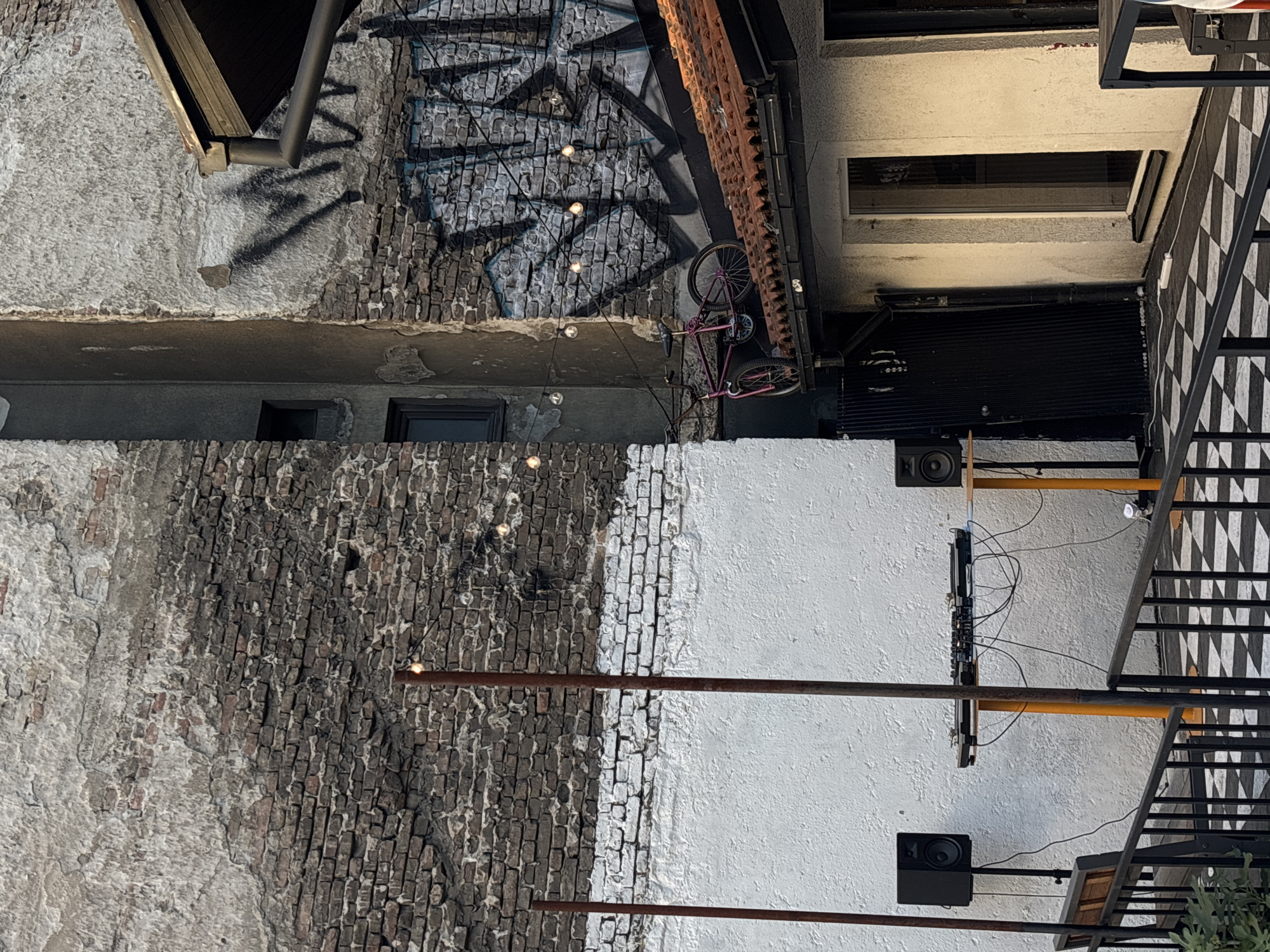 The image depicts an exterior urban scene, seemingly a courtyard or alleyway, in Belgrade, Serbia. The architecture is a mix of old and modern. Walls are constructed from bricks, some covered in white plaster. One wall features what appears to be graffiti or a mural design. A pink bicycle is mounted on one wall. Several metal pipes run horizontally across the scene. Speakers and audio equipment are mounted on the wall. A metal staircase is visible, featuring a black and white checkered pattern. String lights are present.