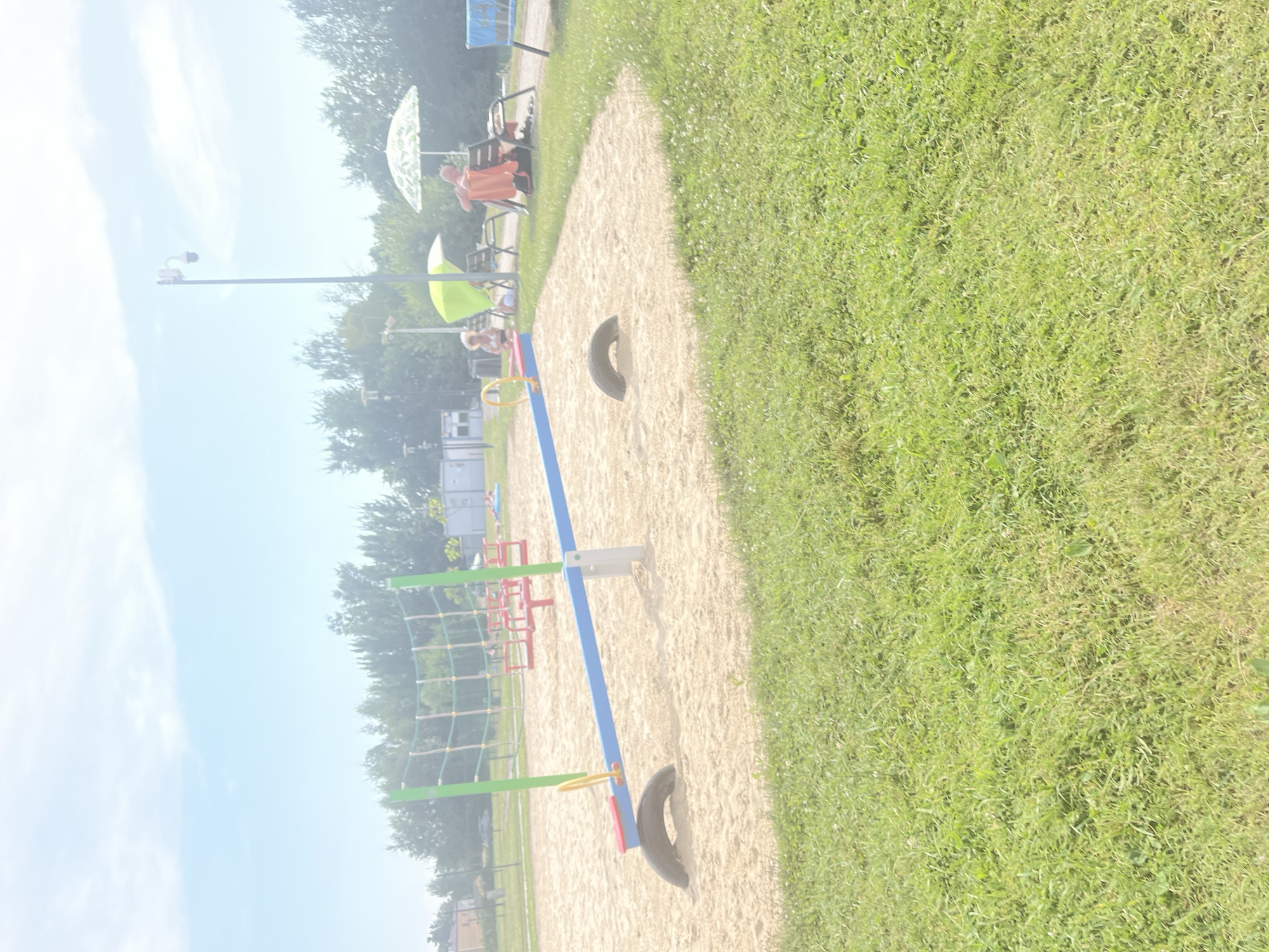 The image captures a sunlit playground scene with sandy ground and green grass. A seesaw, painted blue with red and yellow accents, is a central object. Tires are positioned on the sand, one under the seesaw. In the background, playground equipment with red and green structures and a rope net are visible. Two people are seated on a bench under a large white umbrella, and another person is sitting under a green umbrella. A light pole with a camera is also present. The sky is bright and appears to be clear, suggesting a pleasant, possibly warm day. The image is oriented vertically, with the sky and trees on the left and the playground and grass on the right. There is no visible text in the image.