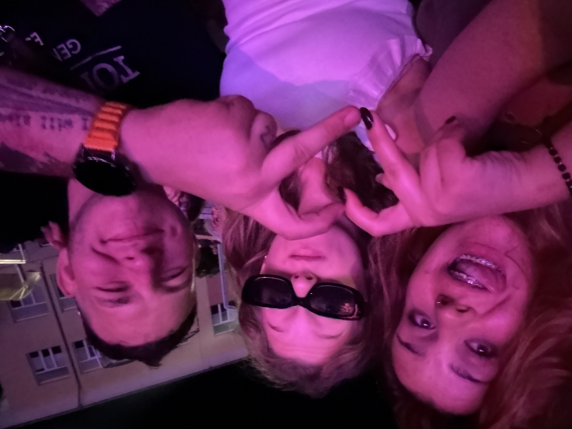 The image shows a close-up of three people, two women and one man, posing for a photo with their hands making heart shapes. The woman on the left has her tongue sticking out and is wearing braces. The woman in the middle is wearing sunglasses. The man on the right is smiling. They are illuminated by pink lighting, suggesting they are at an event or in a dimly lit venue. The background shows a building with windows, indicating an urban or semi-urban environment. The people appear to be in good spirits and are interacting playfully.