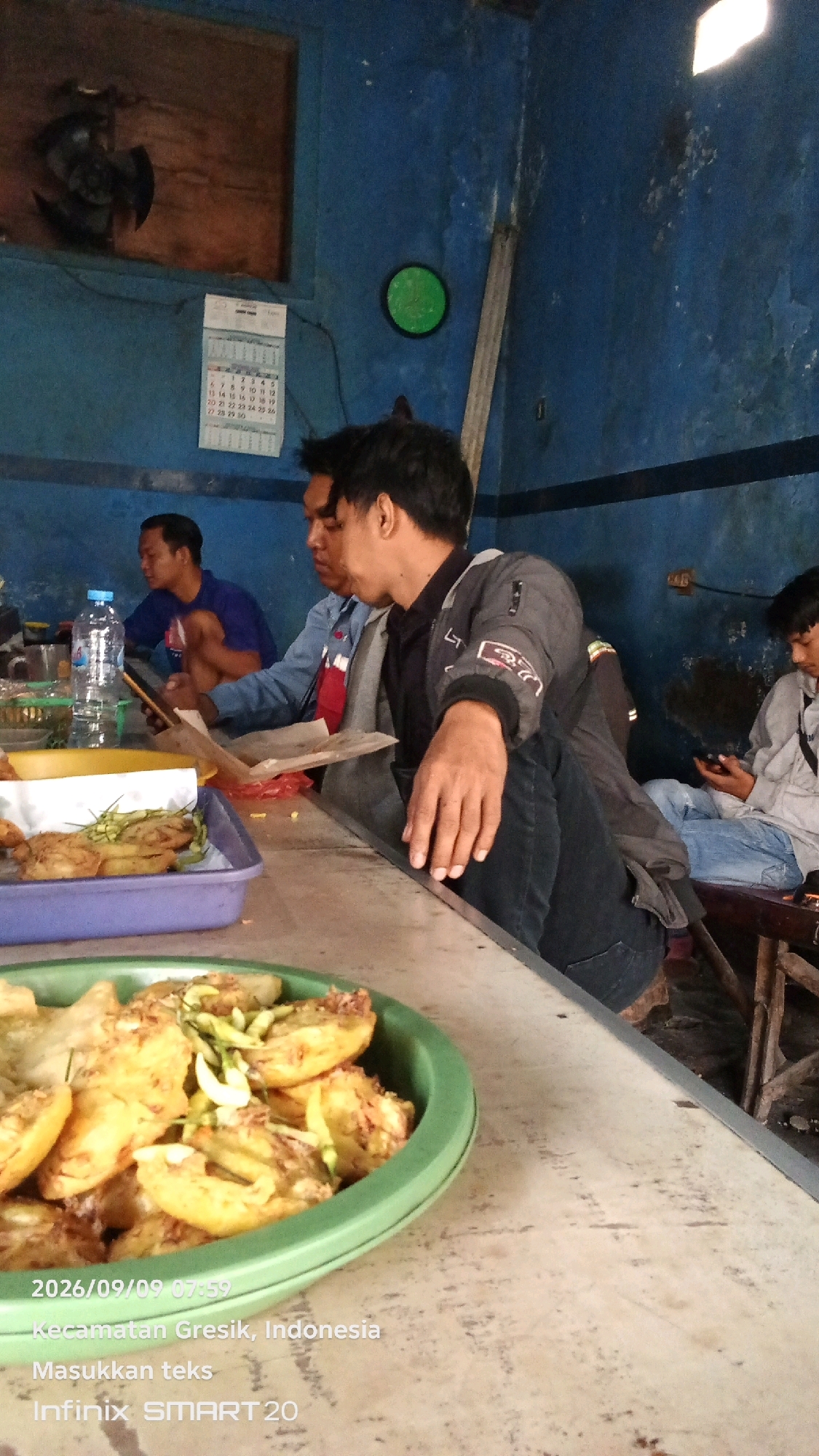 In a dimly lit, blue-walled room, possibly a workshop or casual eatery, four individuals are present. In the foreground, a green bowl filled with fried food, possibly snacks or appetizers, is visible on a table. The food appears to be golden brown and topped with thinly sliced green chilies and what seem to be bean sprouts. The table itself is worn and dusty.
In the mid-ground, three men are seated at the same table. The man on the far left, wearing a blue t-shirt, is looking down at his phone, which he holds in his left hand. Next to him, a man in a blue jacket over a red shirt is also looking at a phone, holding it with his right hand. To his right, a man in a dark jacket with white accents on the sleeves is leaning forward, resting his left hand on the table, his gaze directed downwards. A plastic bottle of water and some paper wrappers are also on the table.
In the background, another person in a light-colored shirt and blue jeans is seated, also looking at their phone. The walls of the room are painted a deep blue and show signs of wear, with some water stains and peeling paint. A calendar with handwritten dates hangs on the wall. A circular green object is mounted on the wall to the left of the calendar. The lighting in the room is artificial and somewhat dim, creating shadows. The overall impression is of a casual, utilitarian space where people are engaged with their phones. The image also includes text overlays indicating the date "2026/09/09 07:59", "Kecamatan Gresik, Indonesia", "Masukkan teks", and "Infinix SMART 20".
