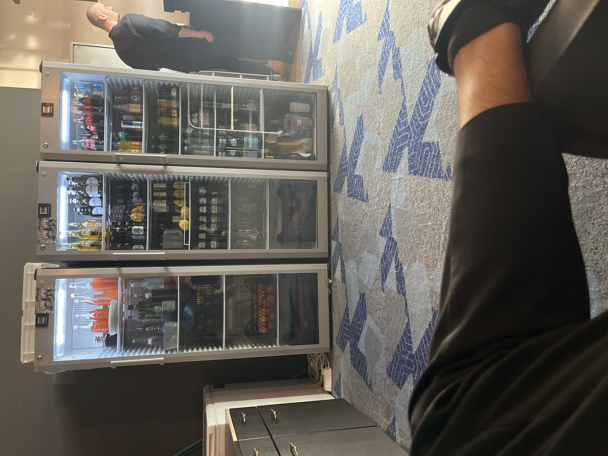 This image captures an indoor scene, likely within a commercial or event setting such as a bar, hotel, or catering area. Three tall, glass-fronted display refrigerators are stacked vertically against a wall.
In the background, a bald man wearing a dark top stands facing away from the camera, looking upwards. In the foreground on the right, the arm and leg of another person, clad in dark clothing, are visible, suggesting they are seated or leaning.
The refrigerators are fully stocked with beverages. The top unit contains various soft drinks, including clearly visible Coca-Cola bottles and cans, and Red Bull cans. The middle refrigerator is filled primarily with beer bottles, many bearing gold-colored labels, and some clear bottles of water or spirits. The bottom fridge holds bottles with orange-colored liquids (possibly juice or cocktails), clear bottled water (including identifiable San Pellegrino bottles), other spirits (such as what appears to be Finlandia or Belvedere vodka), and baskets containing citrus fruit like oranges or lemons.
Below the fridges, a low, dark-colored cabinet with small, gold-toned handles is present, alongside another white appliance or piece of furniture. Electrical cables and a power outlet are also visible. The flooring is a carpet with a light beige/grey background and a repeating blue geometric pattern. The walls are plain and light-colored. The scene is brightly lit by artificial light, particularly within the refrigerators. The overall setup suggests a prepared drinks service area. The location context provided is Sofia, Bulgaria.