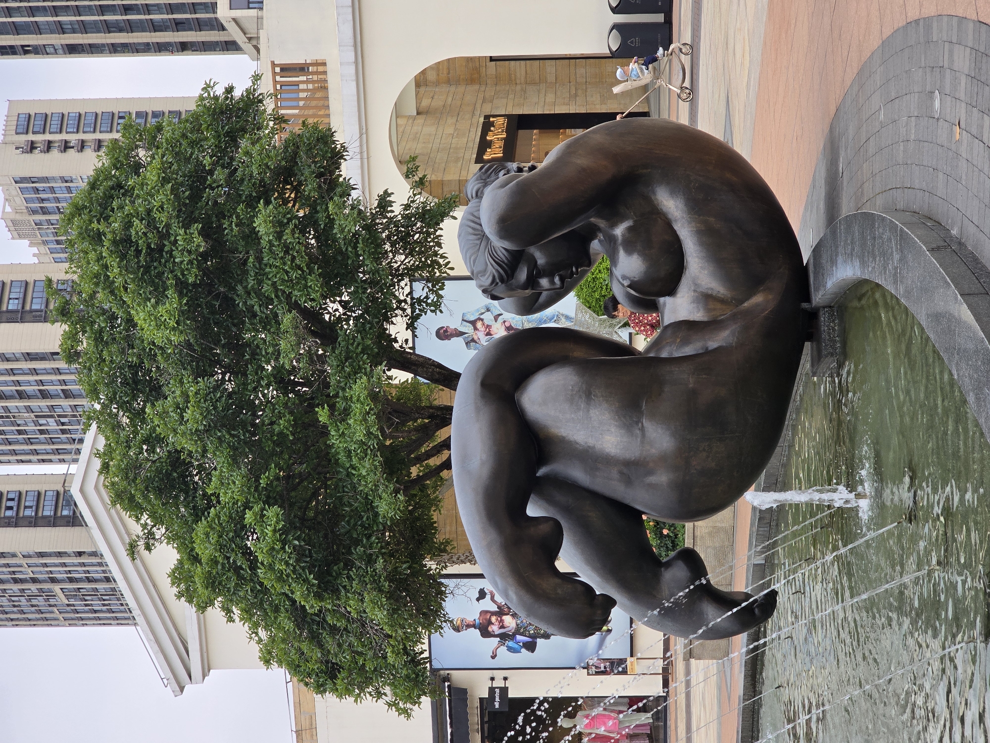 The image shows a large bronze sculpture of a reclining nude female figure in a fountain in Haining, China. The sculpture is situated in a plaza with modern buildings in the background. A large green tree is positioned behind the sculpture. To the left of the sculpture, there are shop windows with mannequins displaying clothing and posters. A fountain with water jets is in the foreground. A child in a stroller is visible on the right side of the image, and other people are in the background. The ground is paved with patterned tiles. The sky is overcast.
