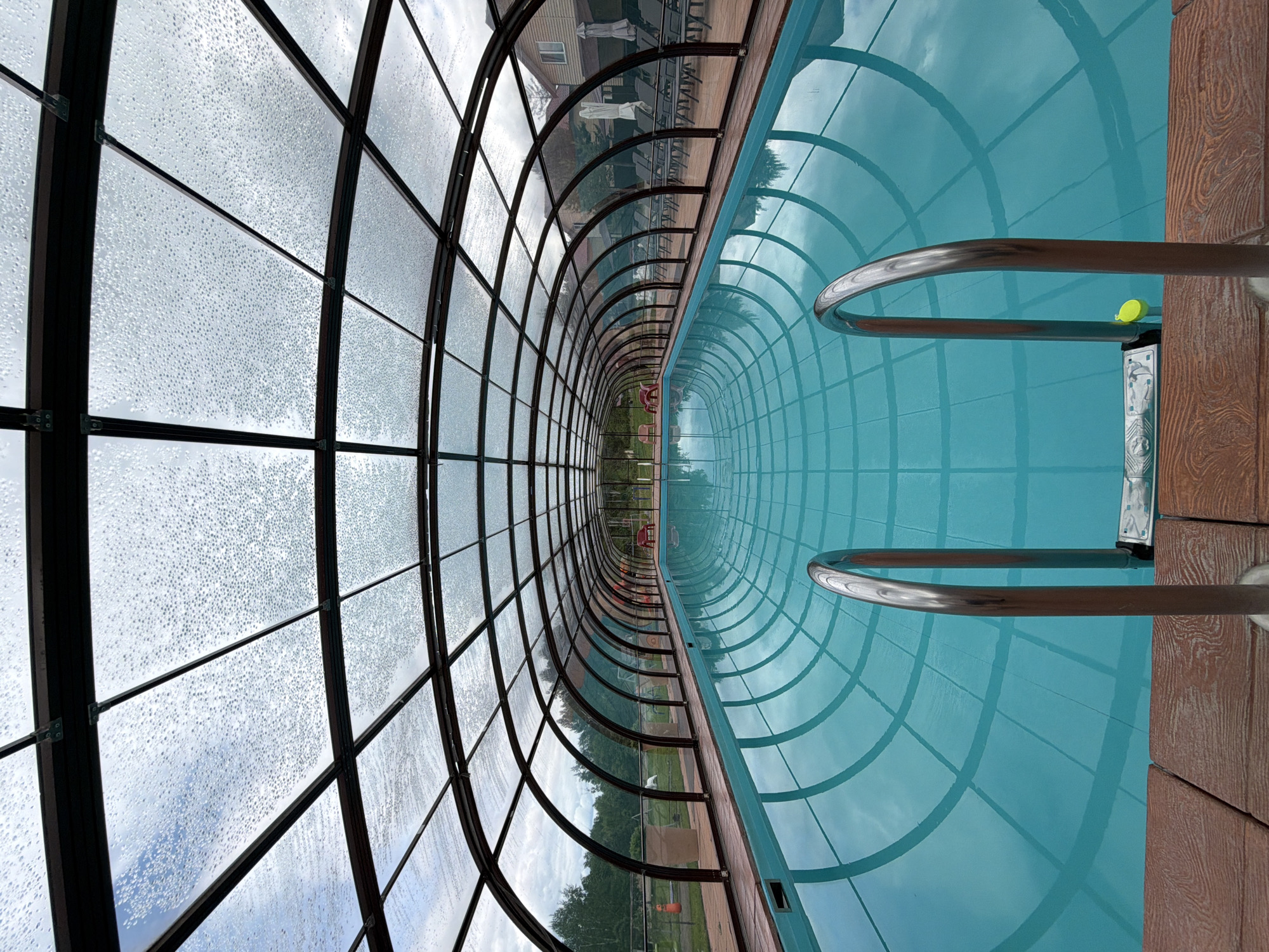 The image shows an enclosed swimming pool with a transparent, arched roof covered in water droplets. The pool is filled with turquoise water and has stainless steel handrails on the right side. The pool deck is made of brown wooden planks. Beyond the pool, lounge chairs and umbrellas are visible, along with some buildings and trees in the background. The scene appears to be outdoors, possibly at a resort or hotel, in a natural setting. There are no people visible in the image. The overall impression is one of a tranquil and somewhat overcast day at an indoor/outdoor swimming facility.
