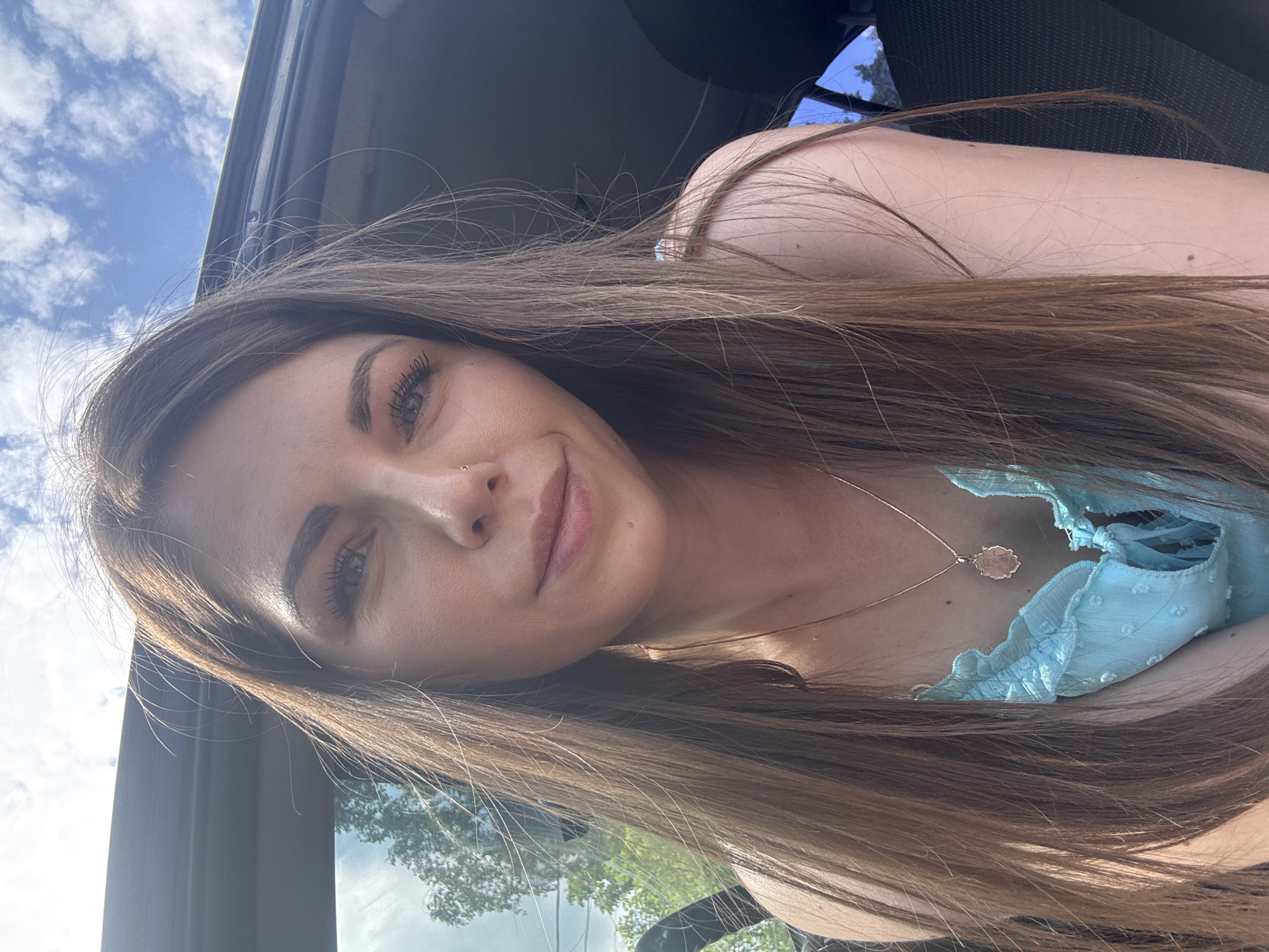 A woman with brown hair, fair skin, and light-colored eyes is pictured inside a car. She is wearing a light blue top and a necklace. The background shows a cloudy sky and trees, suggesting an outdoor setting. No specific location in Shevlyakovo, Russia, is identifiable.