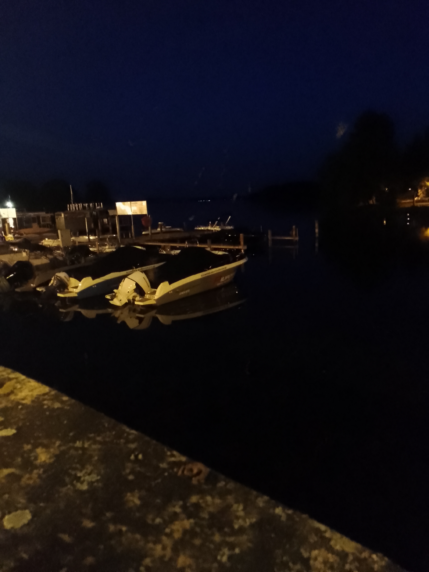 The image depicts a nighttime scene at a harbor in Schwerin, Germany. In the foreground, a stone or concrete embankment is visible, with a dark, textured surface. Beyond the embankment, several motorboats are moored in the water. The boats are covered with dark tarpaulins, obscuring most of their details. Two boats in the center are more prominently visible, with their outboard motors extended. The water is dark and reflects the lights from the boats and the surroundings. In the background, a dock or pier structure with lights is present. Further in the distance, silhouetted trees and buildings are visible, with some scattered lights indicating the presence of a town or city. The sky is a deep, dark blue, suggesting it is late at night. The overall impression is one of a quiet, still harbor after dark.