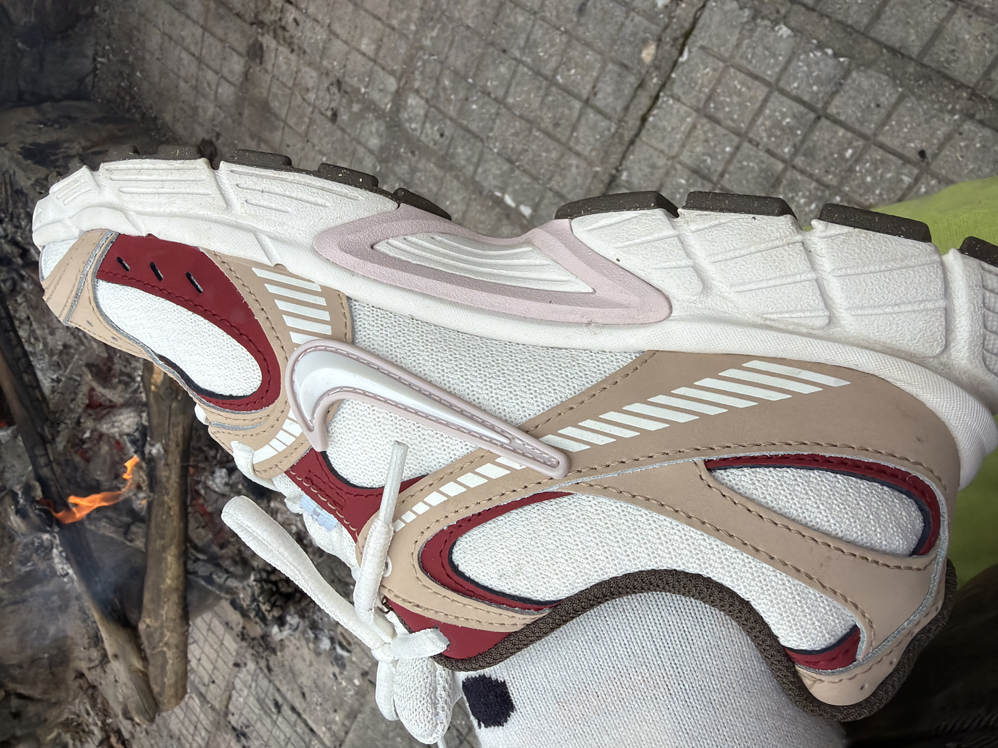 The image shows a close-up of a white, beige, and burgundy running shoe with white laces, worn with a white sock that has a small black spot. The shoe is positioned above a surface of gray stone tiles and a fire pit with visible embers and wood. The location is Lesichovo, Bulgaria, as specified in the prompt.