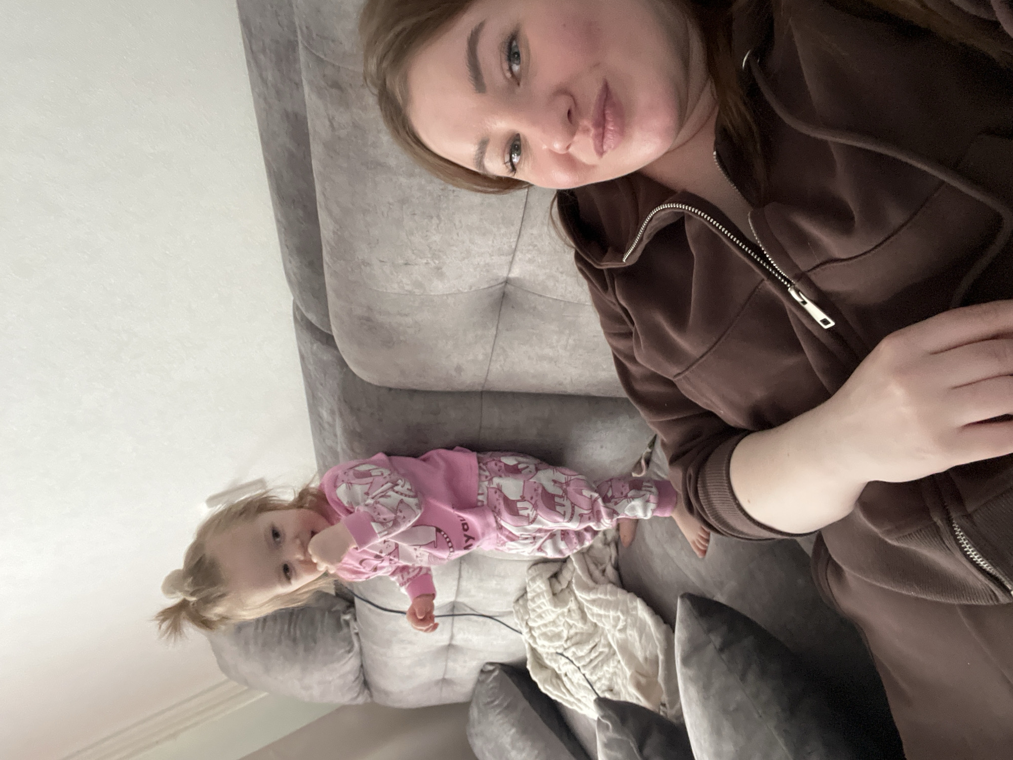 A woman and a young child are on a gray couch. The woman is wearing a brown zip-up sweatshirt. The child is wearing pink pajamas with a cartoon unicorn pattern. The couch has several throw pillows. The photo was taken indoors with a light colored wall in the background. There is no information to suggest this photo was taken in the city of Chaikovsky, Russia.