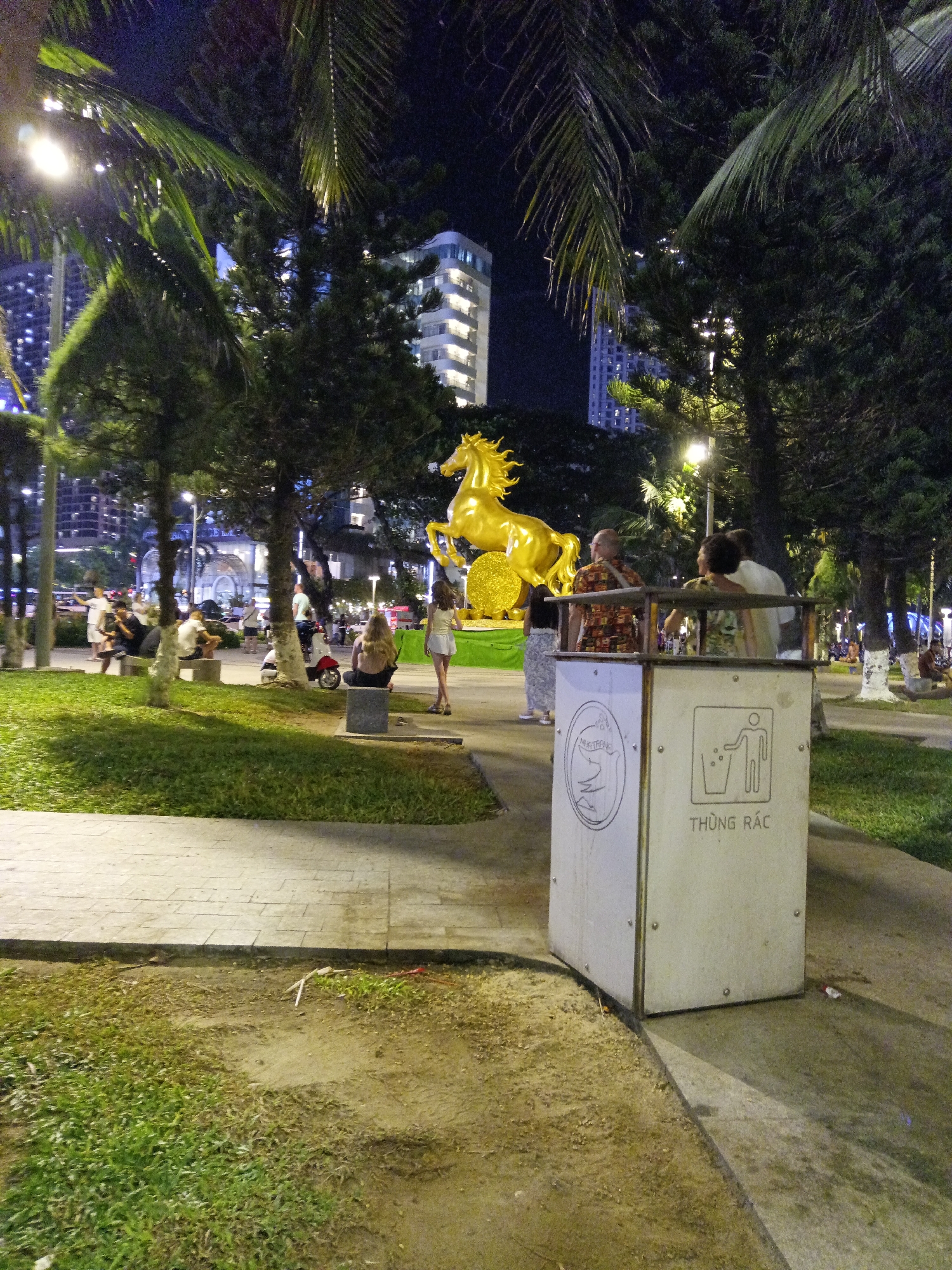 The image depicts a nighttime scene in a park-like area in Nha Trang, Vietnam. A large, golden horse statue is prominently displayed, illuminated by artificial lighting. In the foreground, a white trash receptacle with Vietnamese text "THÙNG RÁC" (meaning trash bin) is visible. Several people are scattered throughout the scene, some sitting on benches and others standing. Trees and palm fronds are present, and tall buildings can be seen in the background, suggesting an urban environment. The ground appears to be a mixture of paved pathways and grassy areas.