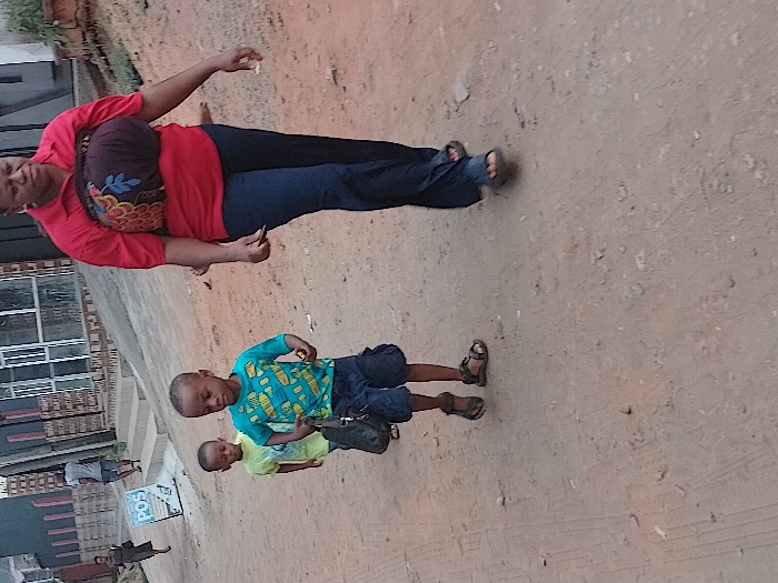 The image depicts an outdoor scene in Orie Uratta, Nigeria, on an unpaved, reddish-brown dirt path. In the foreground, an adult female with dark complexion walks towards the left, wearing a red short-sleeved collared shirt, dark blue trousers, and dark sandals. A dark, patterned fabric with colorful feather and leaf motifs is wrapped around her torso. She holds a small, dark object in her left hand.
Behind her, two young boys follow. The boy closer to the foreground, approximately 5-7 years old, wears a turquoise t-shirt with yellow fish patterns, dark blue shorts, and brown sandals. He carries a black bag in his left hand and a small, light-colored object in his right. The second, younger boy, partially obscured, wears a light green t-shirt and dark shorts.
In the mid-ground, an adult male in a light grey t-shirt and dark shorts walks away from the viewer. Further in the background, another adult female in a dark dress is also walking.
The background consists of several buildings, some with windows, and a set of concrete steps leading up. A white rectangular sign displaying "POS" in blue text is visible on the left side of the path. The ground is uneven, dotted with small stones, and exhibits faint tire tracks. Minimal green vegetation is present on an elevated area to the upper right. All visible individuals are in motion, moving generally from right to left across the frame.