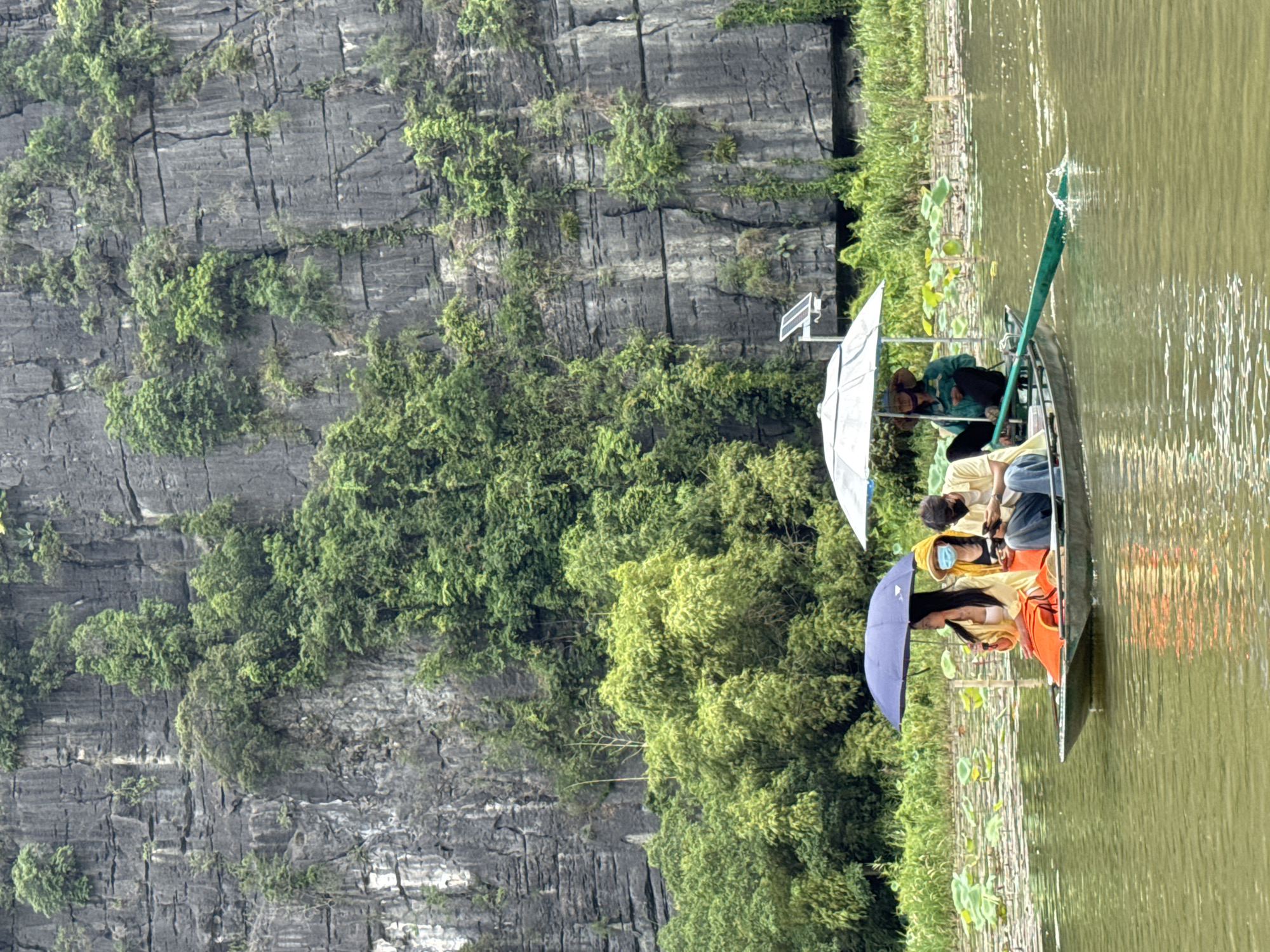 Four individuals are on a narrow boat navigating through a body of water in Nam Hoa Lư Ward, Vietnam. The boat is propelled by an oar, suggesting a leisurely ride. The passengers are partially shaded by umbrellas, one dark blue and another white. In the front of the boat, two women sit side-by-side. One woman, facing the right, wears an orange life vest over a yellow garment and holds a blue umbrella. Next to her, a woman in a yellow hooded jacket and a blue face mask is looking down. Behind them, a man in a yellow shirt and blue jeans, also wearing a black face mask, is looking at his phone. Seated at the stern of the boat, a person wearing a wide-brimmed hat and a green jacket is operating the oar. The environment is characterized by a towering, rocky cliff face covered in lush green vegetation, including bamboo and other trees. The water is calm and reflects the surrounding greenery. Numerous lily pads dot the surface of the water. A small solar panel is visible on the rocky cliff in the background.