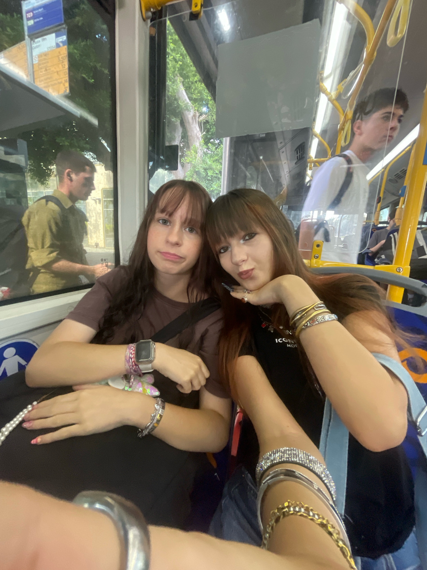 Two young women are seated on a public bus in Raanana, Israel. The woman on the left is wearing a brown t-shirt and has a smartwatch and several bracelets on her left wrist. The woman on the right is wearing a black t-shirt and has multiple bracelets on her right arm, including one with rhinestones. A man wearing a white t-shirt is visible in the background, standing and holding onto a yellow pole. Another man in a green uniform is visible through the window to the left. The bus interior is visible, with yellow handrails and blue seating. Trees and foliage are visible through the windows, indicating an outdoor setting. The overall scene depicts a moment of travel on public transportation.