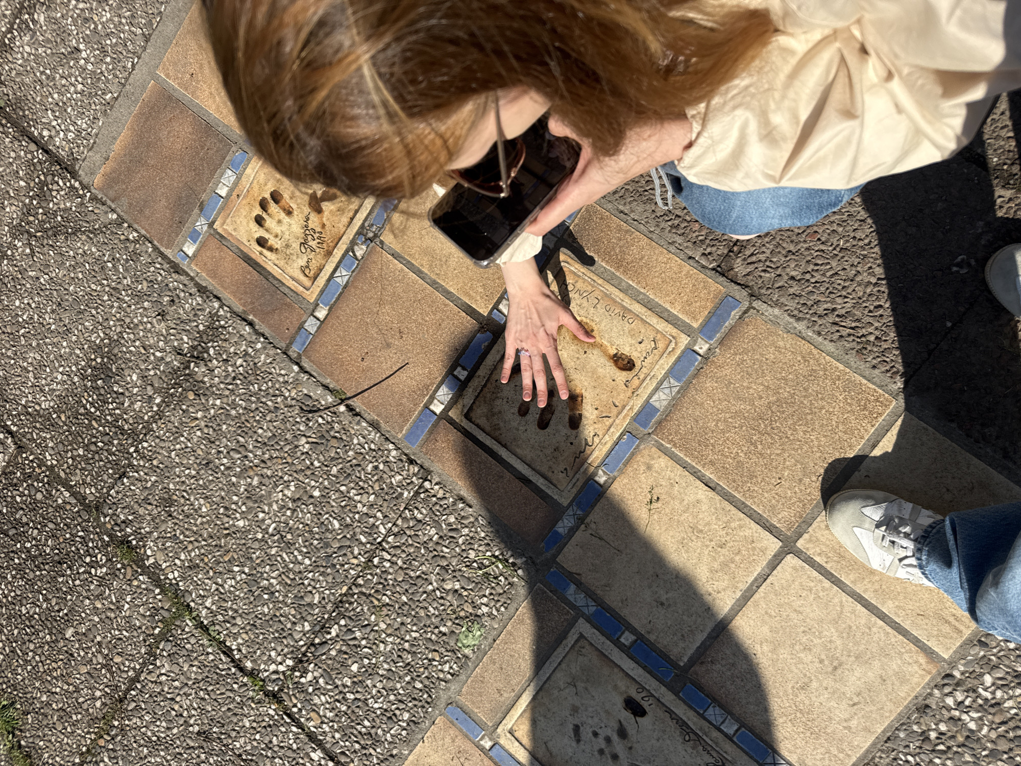 An overhead shot captures a person, likely female, engaged with a celebrity handprint walkway under bright sunlight, indicative of midday or early afternoon. The person, with long, light brown hair, is leaning down, wearing sunglasses pushed up on her head, a light beige jacket, and blue jeans. Her left hand, adorned with a ring, rests flat on one of the square, tan-colored tiles embedded in the ground, seemingly comparing her hand to an imprint or examining the texture. In her right hand, she holds a black smartphone, possibly taking a picture or recording.
The walkway consists of these decorative tiles, framed by blue mosaic borders, interspersed with sections of grey, pebble-dash paving. Numerous strong shadows are cast across the ground, including the person's shadow and the shadow of the smartphone.
Several tiles feature imprints and signatures. Visible text includes "Ben Gazzara" and "1996" next to distinct paw-like impressions on one tile, and "DAVID LYNCH" and "1998" on another. A partial "1990" is also discernible on a third tile. The scene strongly suggests the "Chemin des Étoiles" (Path of Stars) in Cannes, France, known for celebrity handprints. A white sneaker and blue-jeaned leg are visible on the right side of the frame, belonging to another individual or the same person.