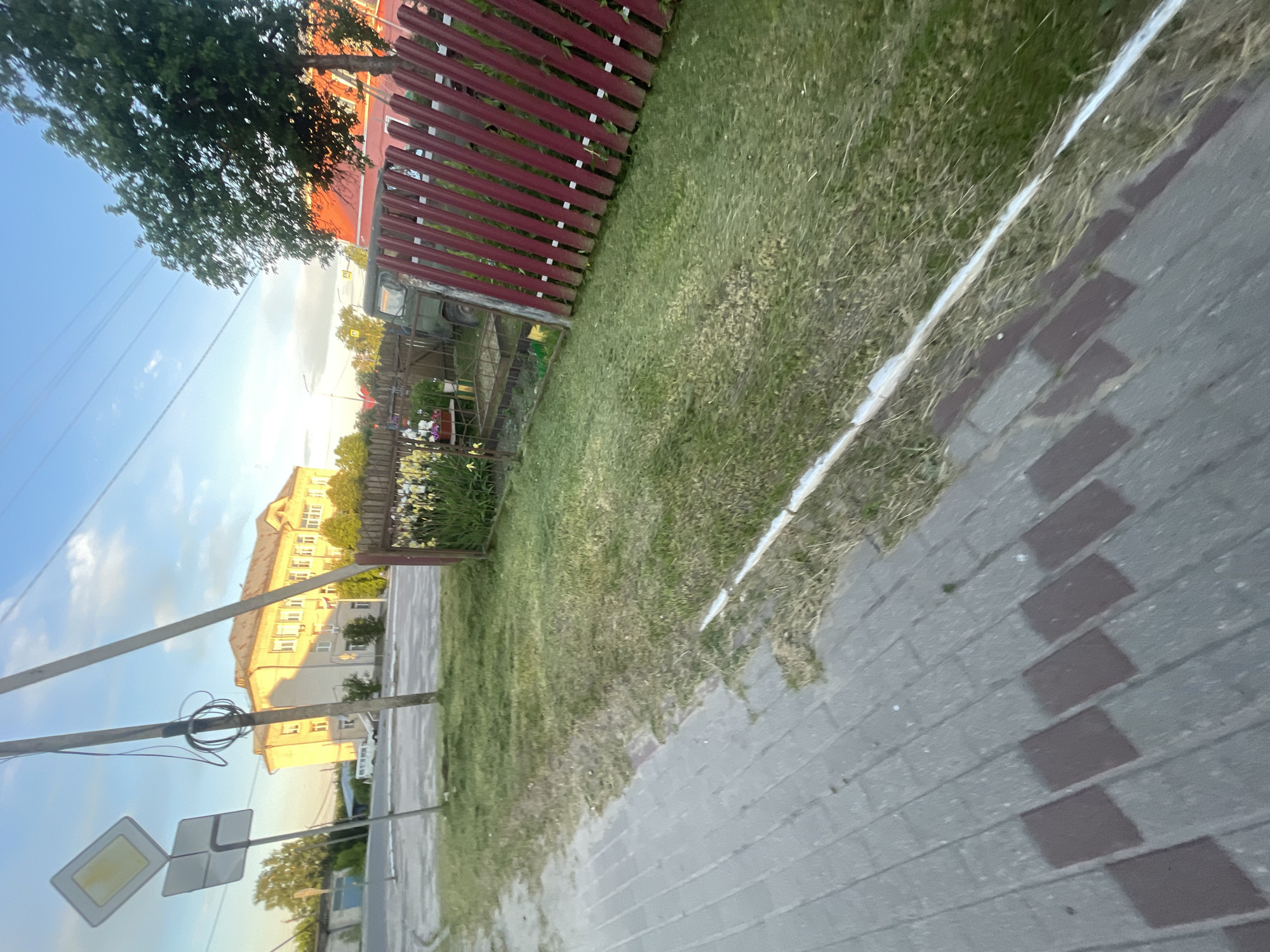 This image shows a sidewalk made of gray and red bricks, with a strip of mown grass adjacent to it, bordering a red metal fence. Beyond the fence, there is a patch of green grass with yellow and white flowers. Further back, a military vehicle is visible, and behind it, a large yellow building stands under a blue sky with clouds. A utility pole with wires and traffic signs is also present in the scene, situated near the sidewalk and the yellow building. The environment suggests a residential or public area within the city of Krasnye Baki, Russia, possibly near a school or government building given the presence of the large structure.