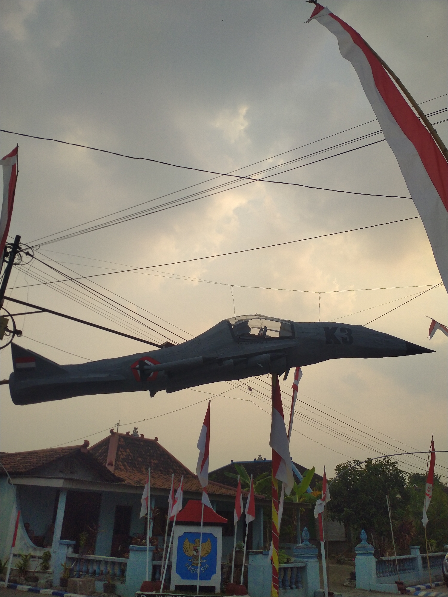 A low-angle, wide shot captures a model of a gray jet fighter plane suspended in the air over a street in Pondok, Indonesia. The plane, which appears to be made of lightweight material, is dark gray with red and white markings and the letters "K3" on its side. Indonesian flags, red and white, are prominently displayed throughout the scene. One large flag is mounted on a bamboo pole on the right side of the image, its fabric billowing in the wind. Smaller flags are attached to poles in front of a building on the left, which is a light blue, single-story structure with a terracotta tiled roof. A coat of arms featuring a golden eagle and the word "INDONESIA" is visible on a blue and white pillar in front of the building. The sky is overcast with diffused light, and multiple electrical wires stretch across the frame. The overall impression is one of a festive or commemorative display, possibly related to a national holiday or event.