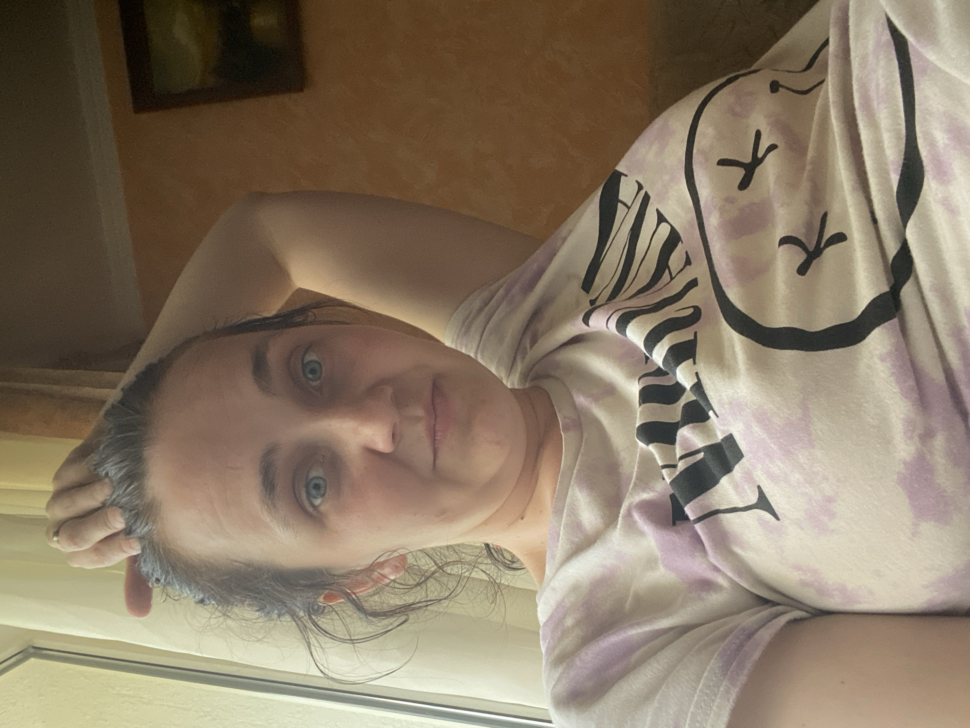 A woman with light blue eyes, brown hair, and fair skin is shown from the chest up. She is wearing a t-shirt with a tie-dye pattern of purple and white. The t-shirt has a graphic design of a face with closed eyes and small 'x' marks for pupils. Her left arm is bent and resting on her head, with her hand touching her hair. The background includes a window with sheer curtains on the left, and a wall with a framed painting on the right. The lighting suggests it is daytime.