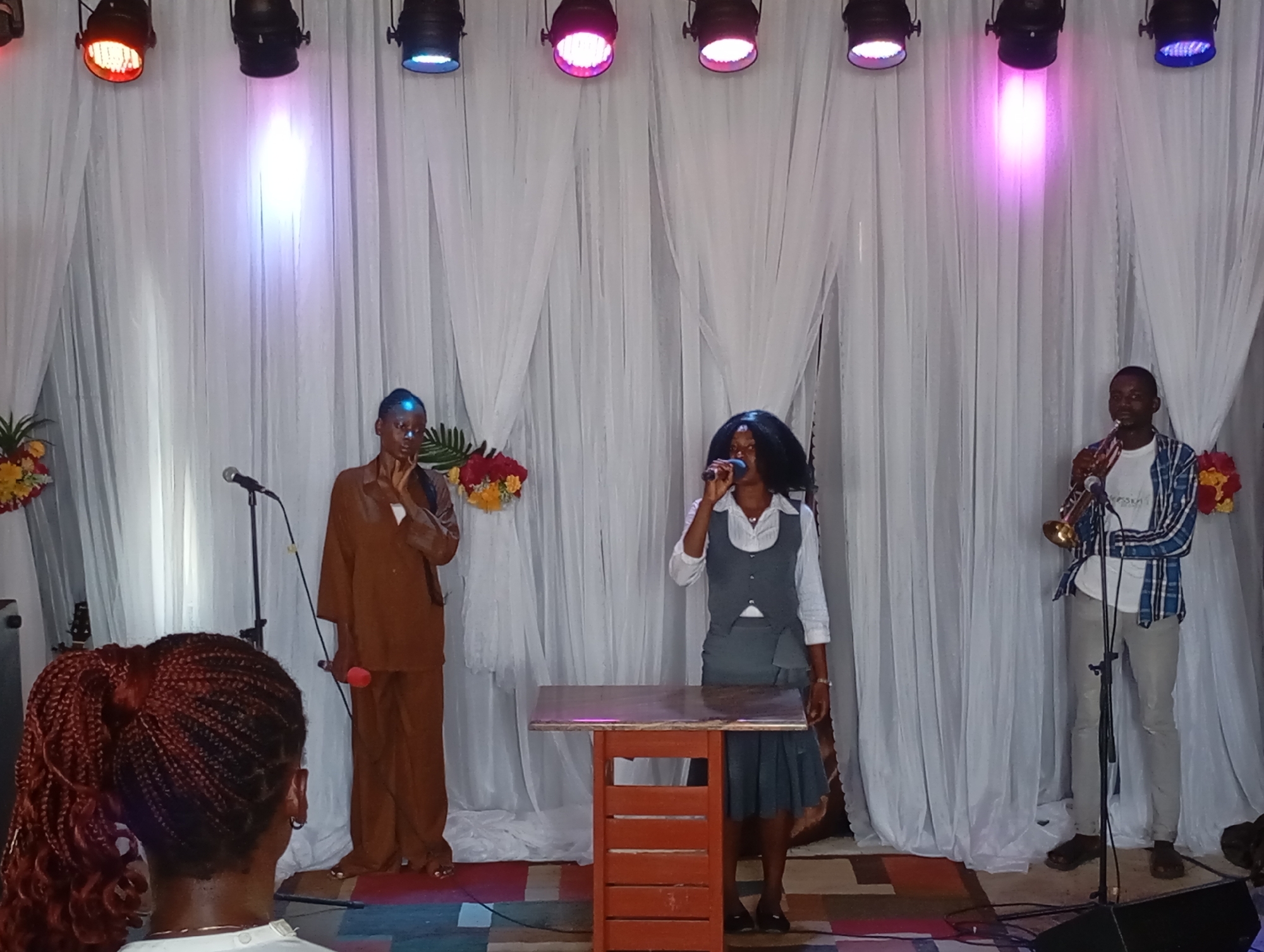 Scene: A stage in Uke, Nigeria.
People: Three performers are on stage. From left to right: A person in a brown jumpsuit holding a red microphone. A person in a white shirt, grey vest and skirt, singing into a microphone behind a small wooden podium. A person playing a trumpet wearing a white shirt and a blue plaid shirt.
Objects: Microphones, microphone stands, a wooden podium, trumpet, stage lights, floral arrangements.
Environment: A backdrop of white fabric draped like curtains. The stage floor is covered in a multicolored geometric pattern. The lights above shine in red, blue and pink hues. A person with braided hair stands in the foreground, facing the stage.