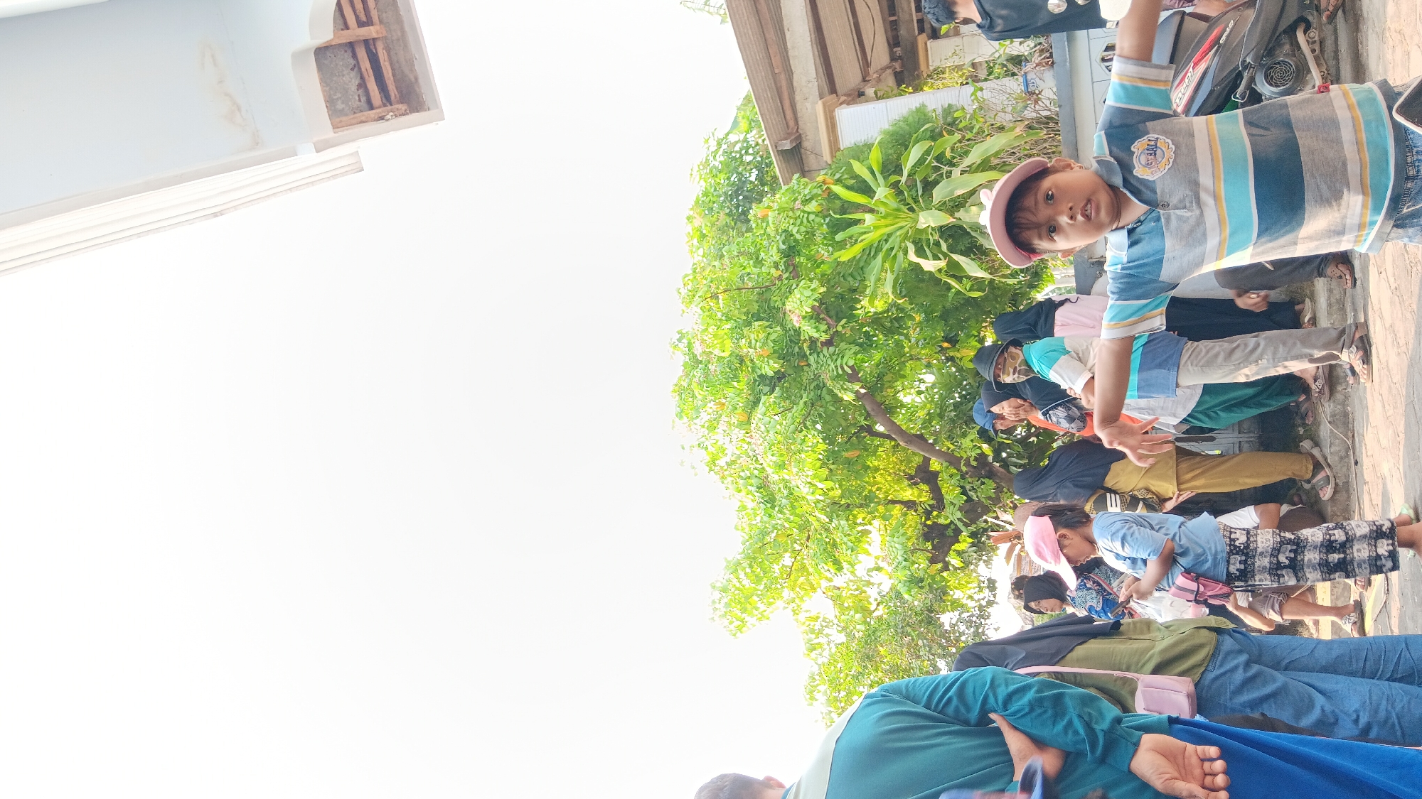 In the city of Cerme, Indonesia, a group of people are gathered outdoors, likely on a street or pathway. The scene is dominated by a large tree with vibrant green foliage. In the foreground, a young boy wearing a striped polo shirt and a pink cap is looking towards the camera with his arms outstretched. Behind him, other individuals, including women wearing hijabs, are visible, some appearing to be queuing. The background features a building with a portion of a roof and some wooden elements. The overall atmosphere seems to be that of a casual outdoor gathering.