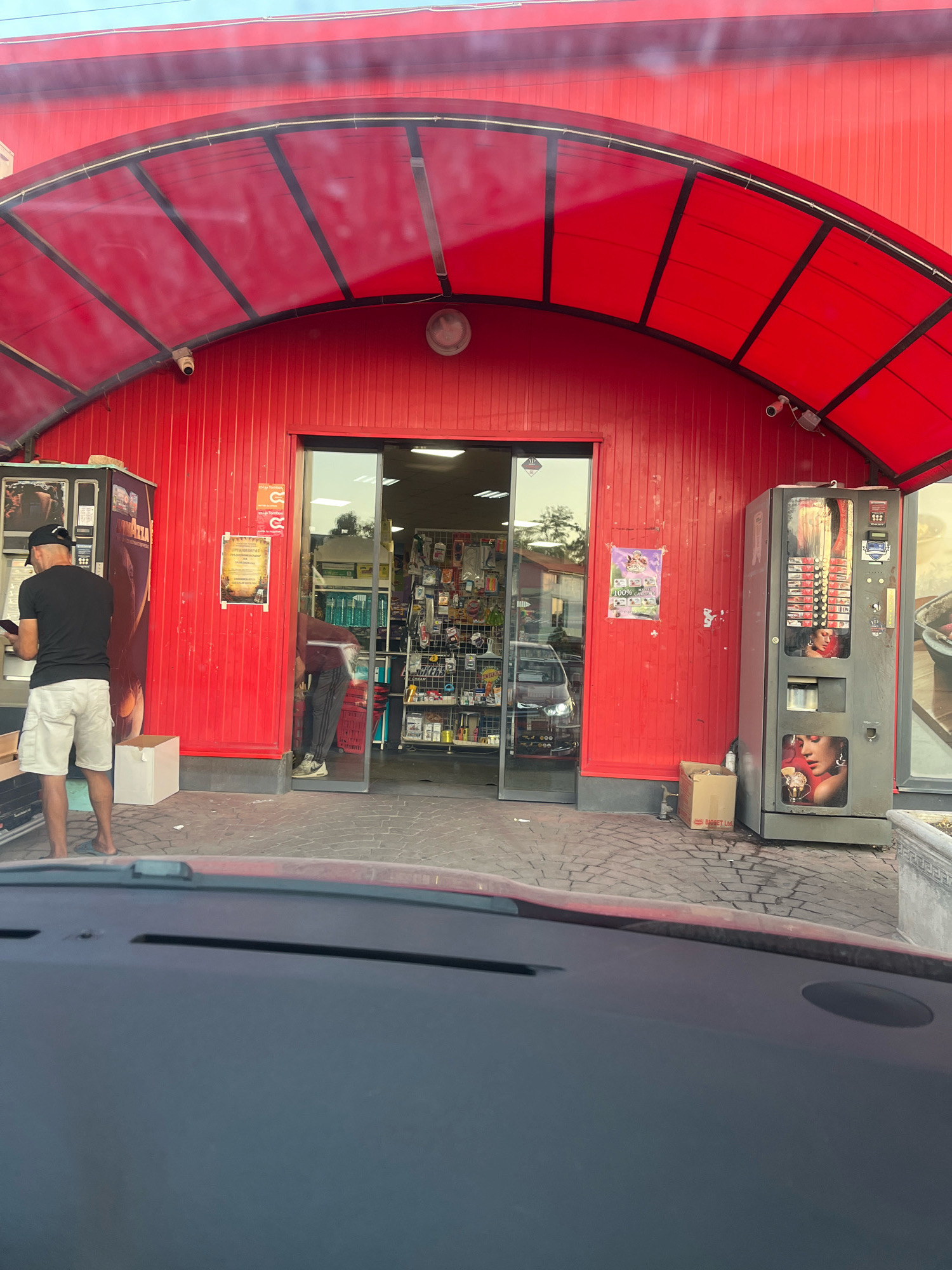 A man wearing a black t-shirt, white shorts, and a black baseball cap is standing outside a red building with a red awning. He is looking at a mobile phone in his hands. To his left is a vending machine. The building appears to be a shop or convenience store, with a glass-fronted entrance in the center. Inside, shelves are stocked with various products. Another person, wearing a maroon t-shirt and dark pants, is visible inside the store. There is another vending machine to the right of the entrance. The ground is paved with interlocking stones. The scene is outdoors, with a clear sky. The location is the city of Sadovo, Bulgaria.