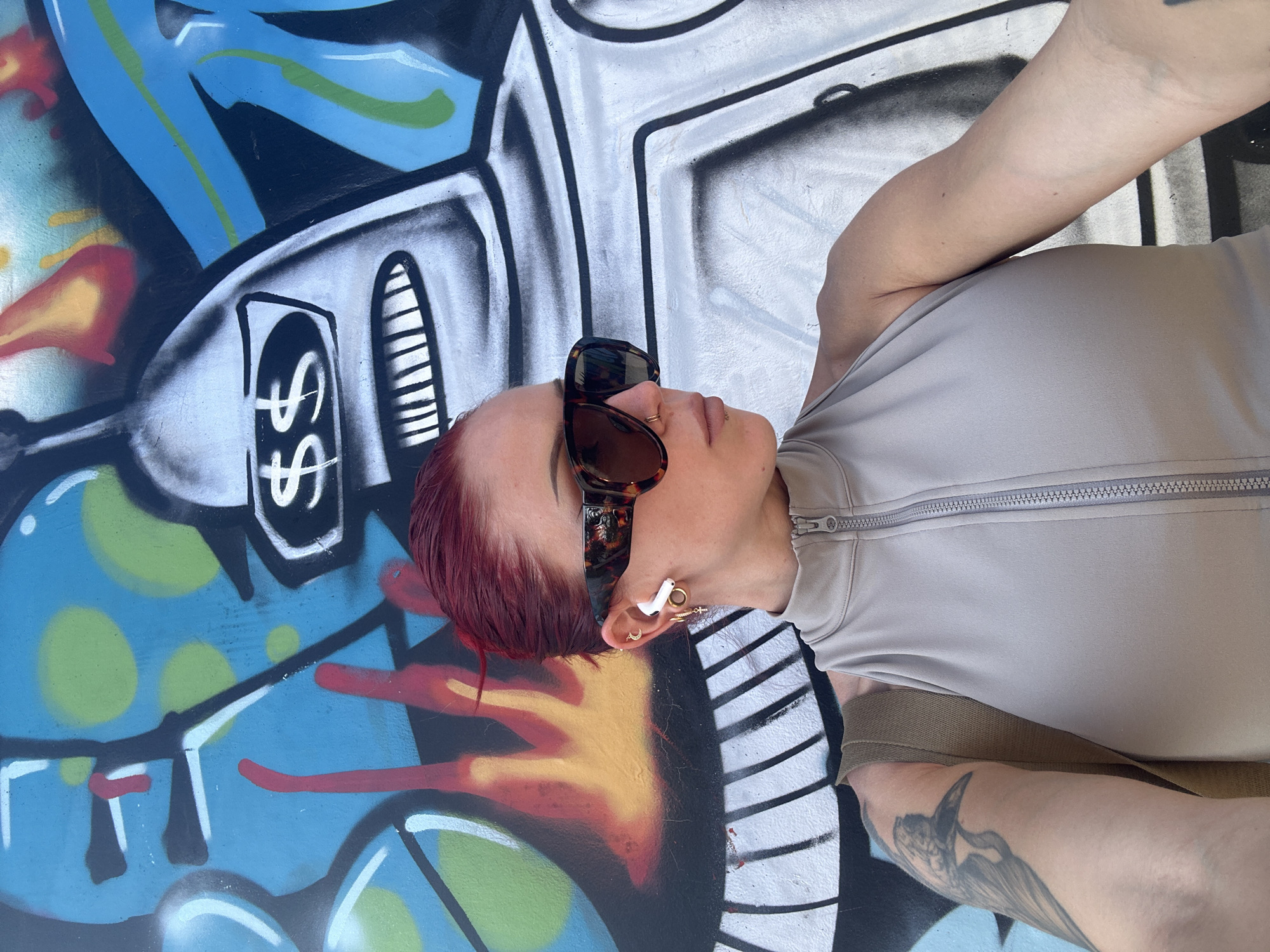 A person with reddish-purple hair and sunglasses lies in front of a colorful graffiti mural. The person wears a zippered top and appears to have tattoos on their arms. They also have several piercings in their ear. The mural behind them contains elements of blue, white, black, and yellow paint. There is no verifiable evidence in the image to definitively place the scene in North Kuta, Indonesia.
