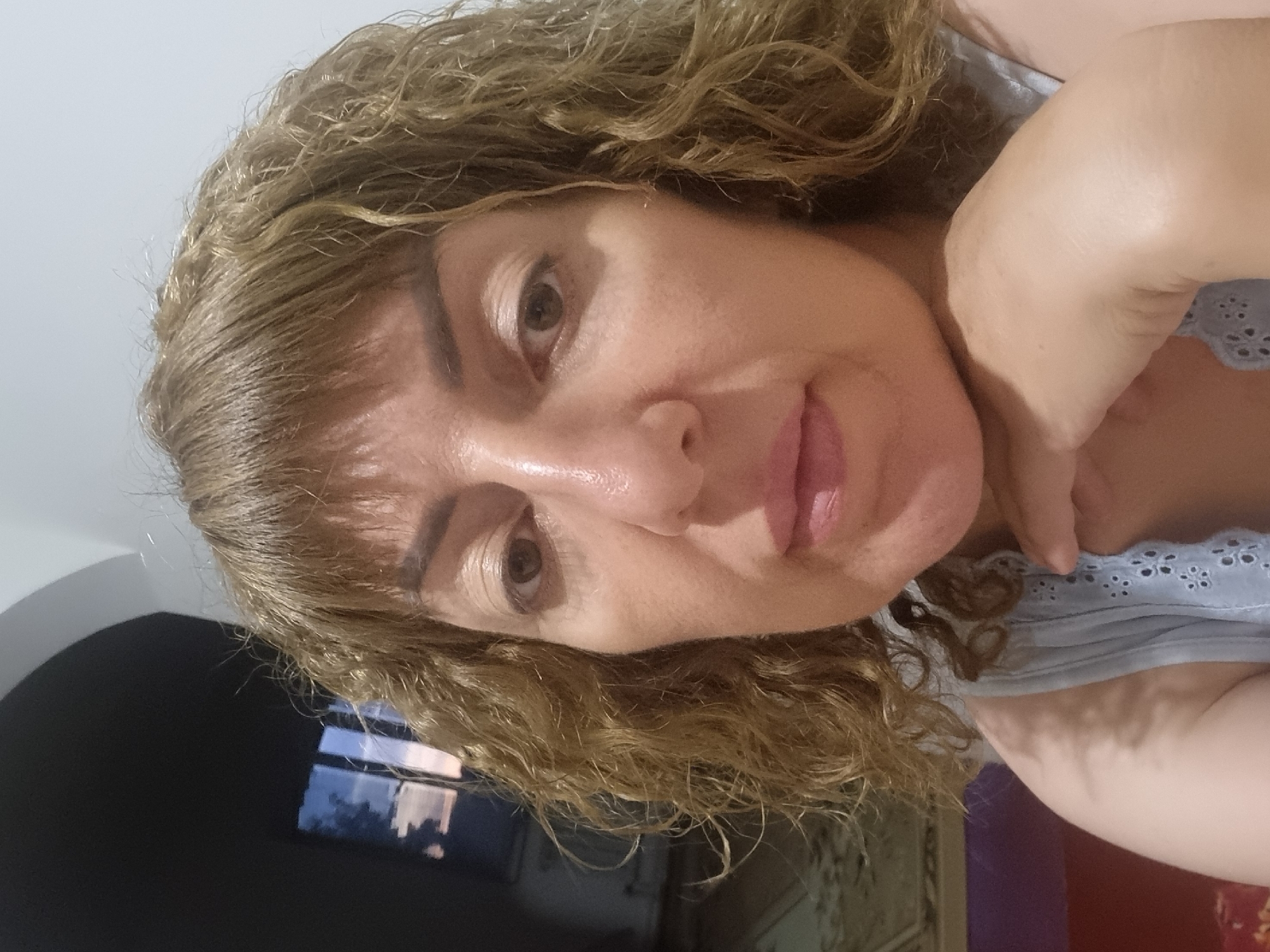 A medium shot photograph shows a woman with shoulder-length, curly, light brown hair and bangs. She is looking directly at the camera with her chin resting on her left hand. The woman has light-colored skin, brown eyes, and a neutral expression. She is wearing a light blue top with an eyelet detail around the neckline. The background is dimly lit, with a window on the left showing a twilight sky. The setting appears to be indoors, possibly a residential interior. There is no visible indication of the specific location within Blizhnee, Ukraine.