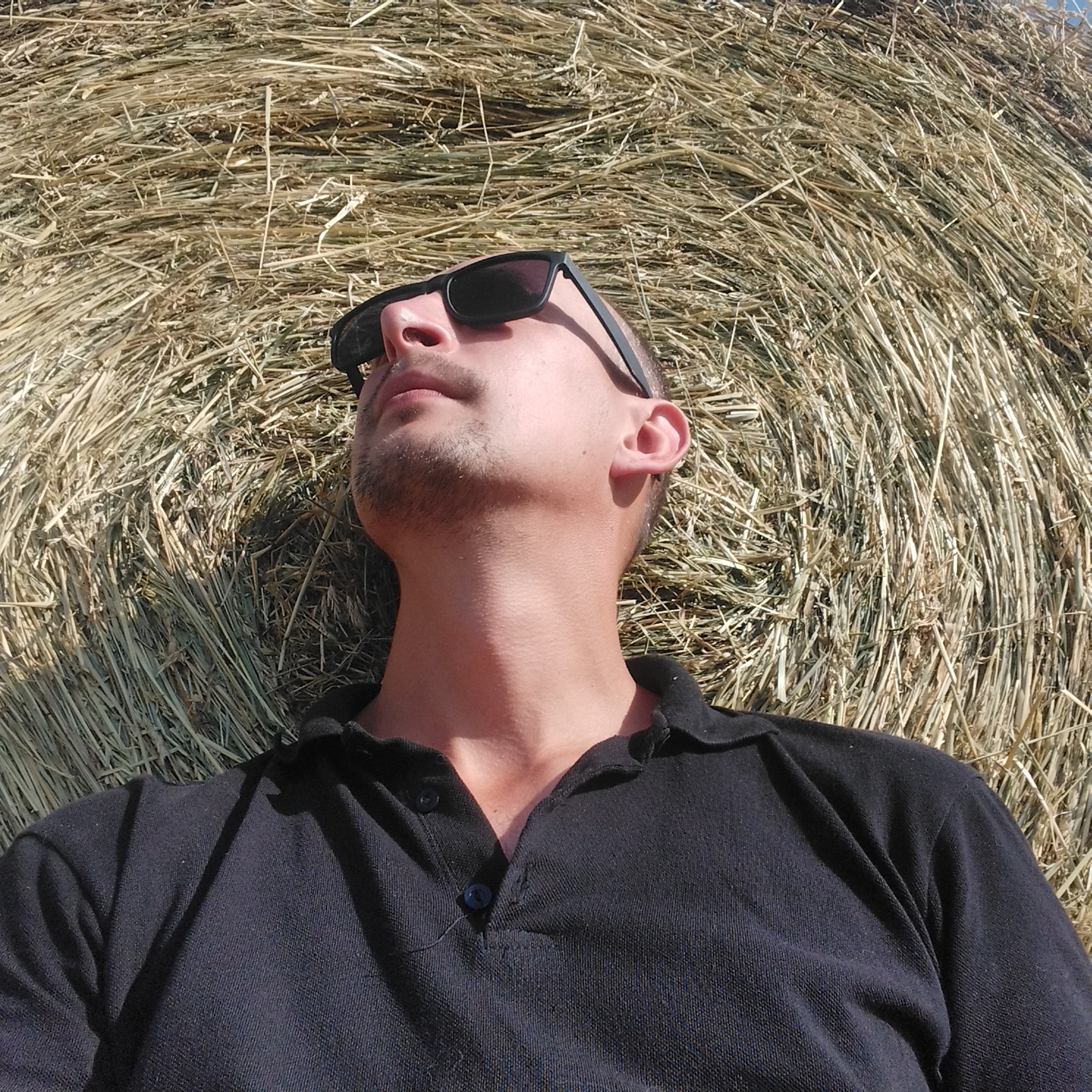 A medium shot, slightly high-angle, indoor portrait of a light-skinned male with stubble, wearing black sunglasses and a black polo shirt, lies on his back against a large round hay bale. The hay bale fills the background with a textured pattern of dried grass stalks. The man's head is tilted back and his face is angled slightly upward, looking towards the top of the frame. Sunlight casts shadows across his face and the hay. The man's eyes are obscured by the dark lenses of his sunglasses. His lips are slightly parted. A few strands of dark hair are visible at the sides of his head. The black polo shirt has a collar and two visible buttons at the neckline, which is unbuttoned. One dark button is visible in the center of the placket, and another is visible on the left side. The scene appears to be outdoors on a sunny day, based on the lighting and shadows. The location is unspecified but implied to be a rural or agricultural setting.
