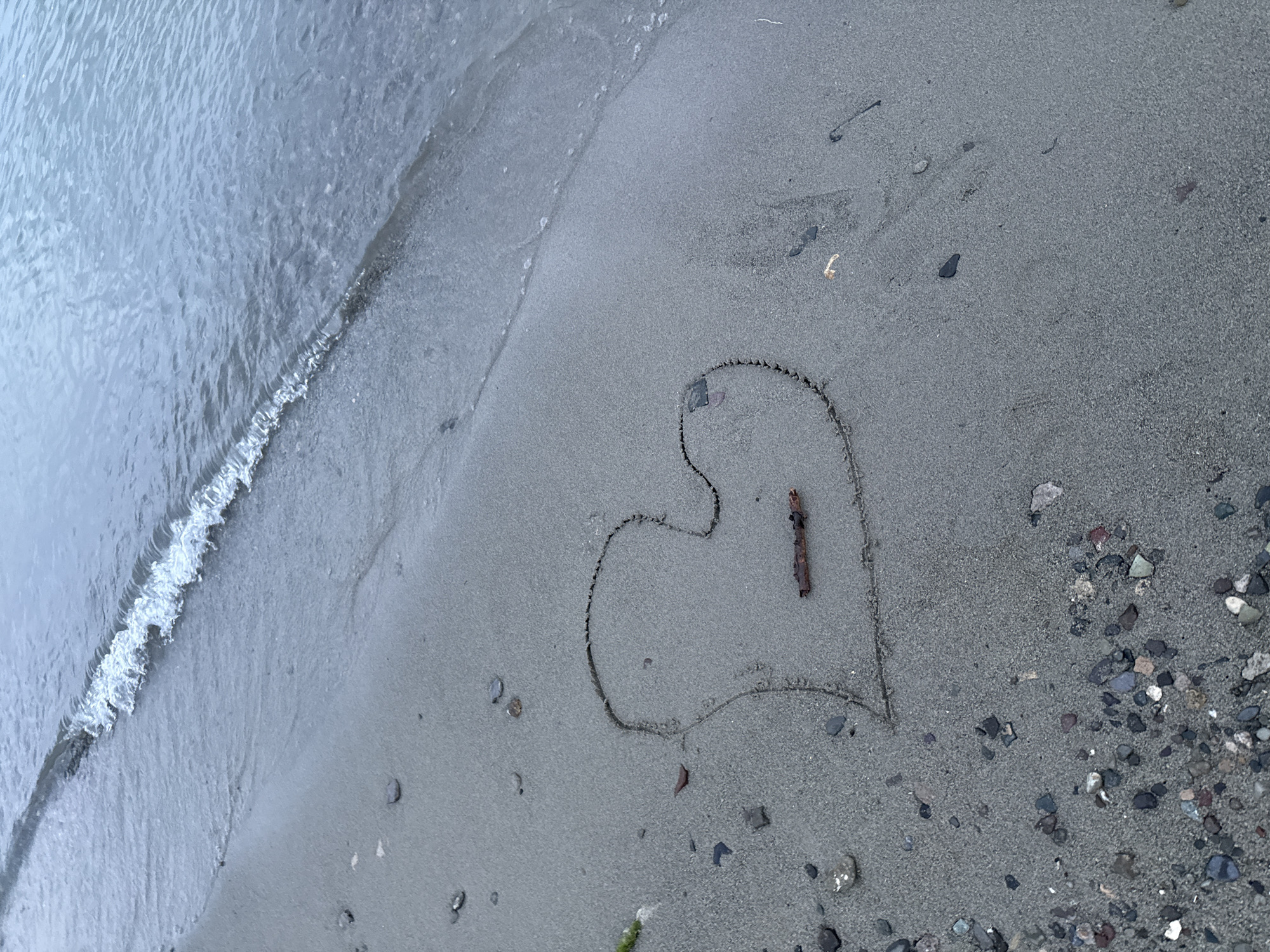 A heart shape is drawn in the wet sand on a beach. A small piece of driftwood is placed horizontally across the middle of the heart. The gentle waves of a body of water lap at the edge of the sand. The sand is a light gray color and is scattered with small stones and pebbles. The water is a calm blue-gray. The image does not provide enough information to determine the location within Tsamakaberd, Armenia.