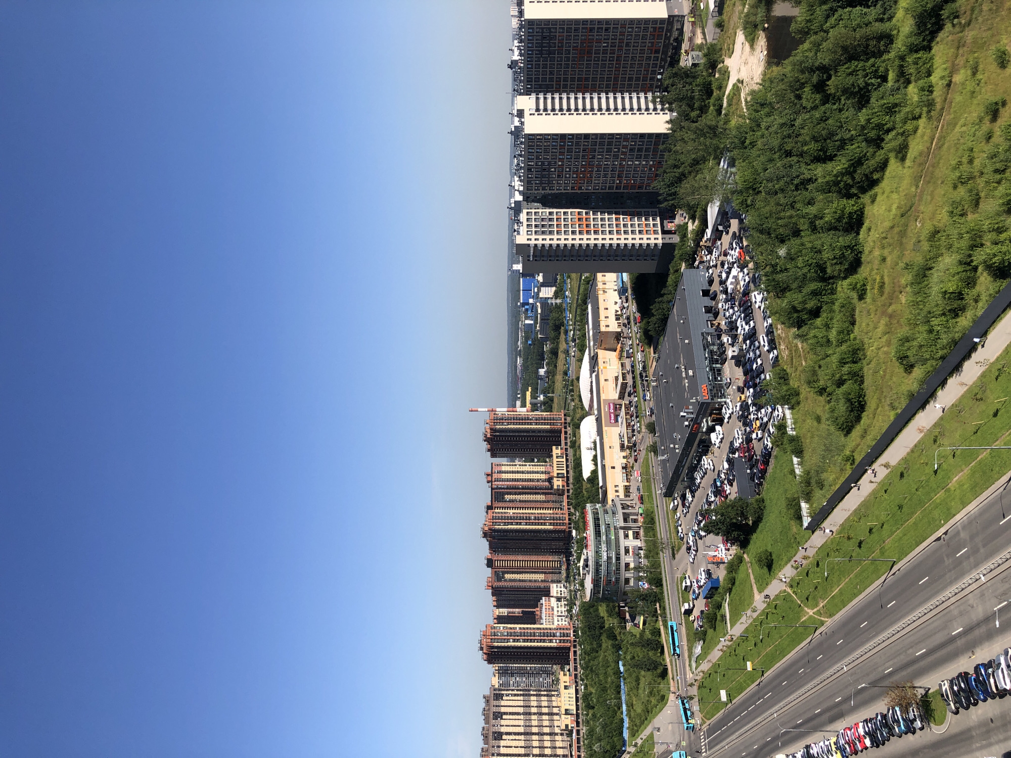 The image captures a daytime aerial view of a suburban area within a city, characterized by modern residential buildings, commercial structures, and infrastructure. In the foreground, a multi-lane road runs diagonally from the bottom left to the mid-left of the frame, lined with parked cars. A pedestrian walkway with lampposts runs parallel to the road, flanked by green grassy areas and trees.
Dominating the mid-ground is a circular commercial building with a sign that reads "TREND CITY." Adjacent to it are large, light-colored commercial buildings with signage, suggesting a retail or entertainment complex. A vast parking lot filled with numerous cars is situated in front of these buildings.
Further back, a cluster of tall, modern residential buildings with reddish-brown facades and numerous windows is visible. To the right, another group of taller, gray and white residential buildings with dark-paned windows stands out. Beyond these structures, the landscape extends to a hazy horizon with more buildings and industrial elements.
The sky is a clear, bright blue, indicating a sunny day. The overall scene depicts a developed urban or peri-urban environment with a mix of housing, commerce, and transportation networks.