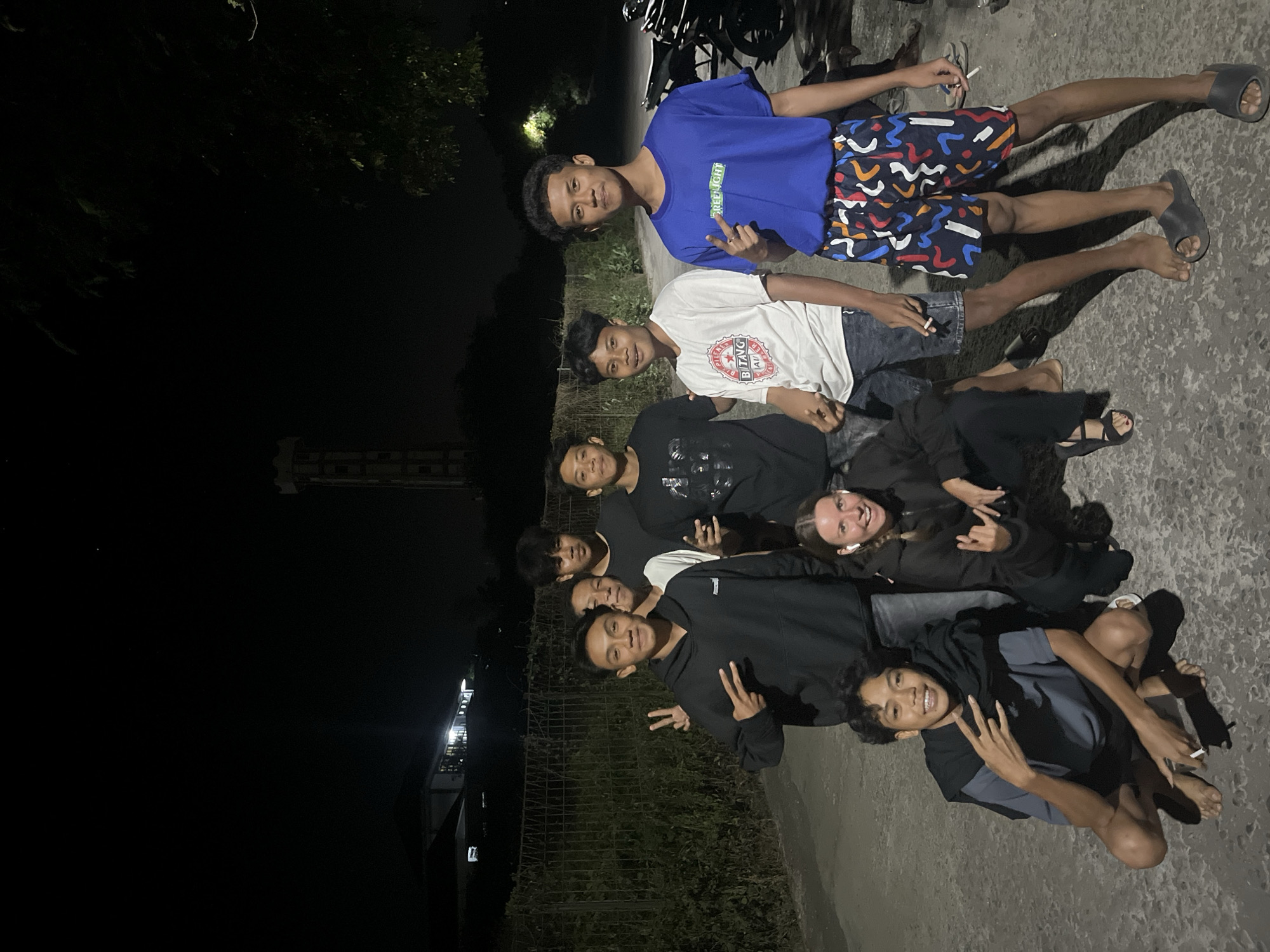 In Labuhan Lombok, Indonesia, a group of eight young people, seven males and one female, are gathered outdoors at night. They are standing and crouching on a paved surface. The individuals are dressed casually, with the males wearing t-shirts and shorts, and the female in a dark hoodie and pants. Two of the males are making peace signs with their fingers. One male in the foreground is crouching and holding a cigarette. In the background, there is a chain-link fence, foliage, a building with lights on, and a tall, illuminated structure that appears to be a water tower. A few motorcycles are parked to the right of the group. The lighting suggests artificial illumination from nearby sources.