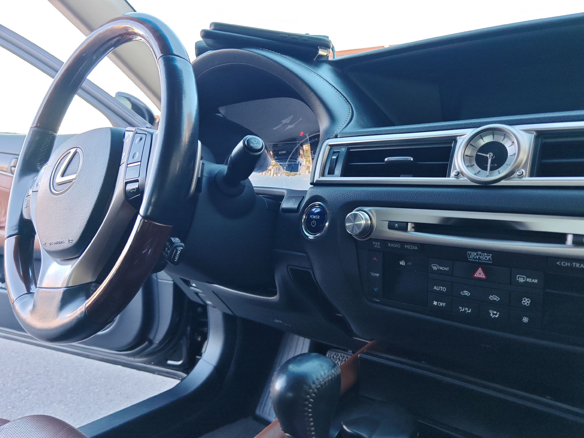 The image captures the luxurious interior of a Lexus car, viewed from the driver's side floor up towards the dashboard and windshield. No people are visible in the scene. The primary subjects are the dark wood and leather-trimmed steering wheel with a "Lexus" logo and control buttons, and the sophisticated dashboard and center console.
The dashboard features an instrument cluster with reflections of a cityscape, and a phone is placed on top. To the right, the center console includes an elegant analog clock with a silver bezel, flanked by air vents. Below this, a multimedia and climate control panel is visible. The gear shifter, wrapped in black stitched leather, is in the foreground at the bottom right. The car's interior features dark materials, including black and brown leather on the seats and trim, contrasted with metallic accents around buttons and vents.
Notable details include the bright daylight illuminating the car's interior, suggesting a clear day. The reflection in the instrument cluster indicates an urban setting, consistent with the provided location context of Sochi, Russia.
Visible text includes:
"SRS-AIRBAG" on the steering wheel, "ON/OFF" on a stalk, "POWER" with a power symbol on a circular button, "PWR VOL" next to a rotary knob, "RADIO MEDIA" above a set of buttons, "Mark Levinson PREMIUM SURROUND" indicating the audio system, "FRONT" and "REAR" on defogger buttons, "TEMP" for temperature controls, "AUTO" and "OFF" for climate control, and partially visible "<CH-TRA" (likely Channel Track) on the infotainment controls. "AUTO HOLD" is also visible near the power button.