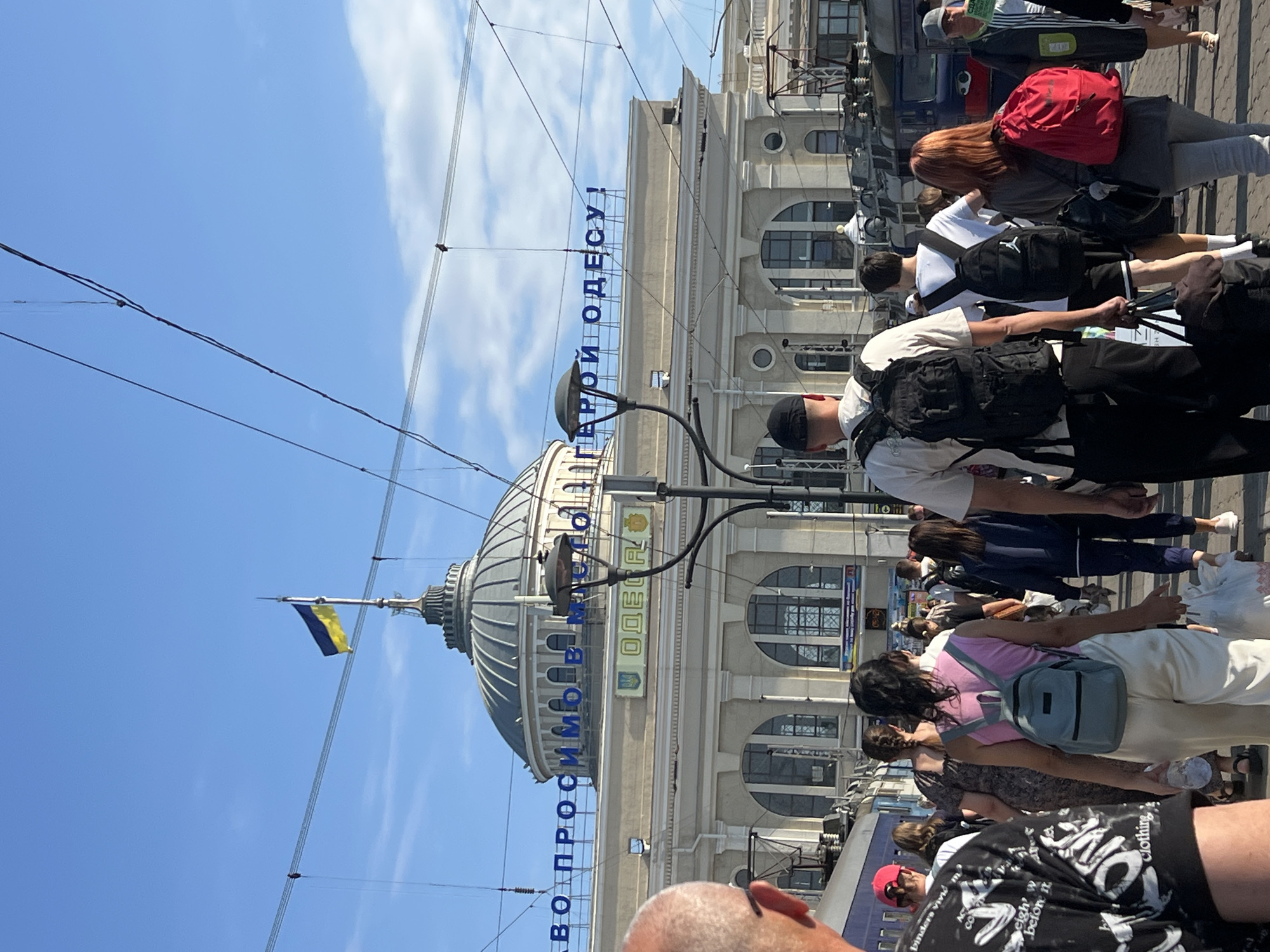 The image depicts the exterior of the Odesa Railway Station in Odesa, Ukraine. A Ukrainian flag flies from the top of the station's dome, which is flanked by the words "ЗАВОД ПРОСИМОВ МІСТО ГЕРОЙ ОДЕСУ!" in large blue letters. The station building is light-colored with arched windows. A crowd of people, many carrying luggage and backpacks, are walking in front of the station. Some are looking towards the station, while others are walking away. The scene is set under a clear blue sky. The overall impression is that of a busy transportation hub.