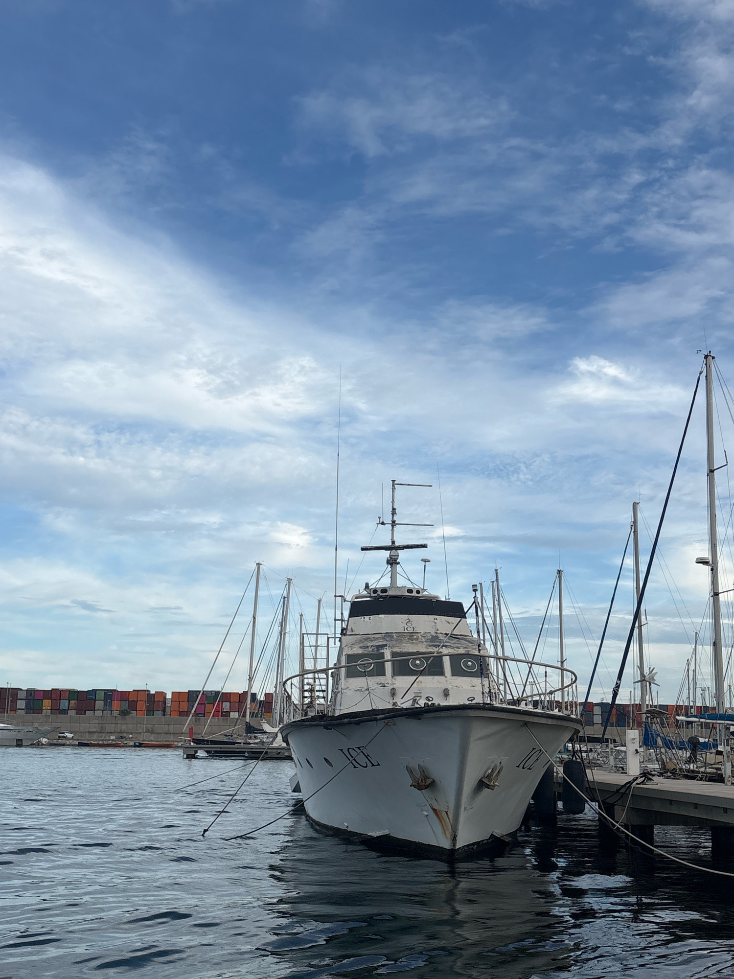 A white vessel, likely a tugboat, with "ICE" written on its bow, is moored at a dock in a marina. The boat has multiple windows and a radar antenna. Several other sailboats and yachts are docked nearby, their masts visible against a blue sky with scattered clouds. In the background, a row of colorful shipping containers is stacked along the waterfront. The water in the foreground is rippled and reflects the sky and the boats. The scene suggests a sunny day in a port city. This is likely the Port of Valencia, Spain, given the presence of shipping containers and the type of vessels.
