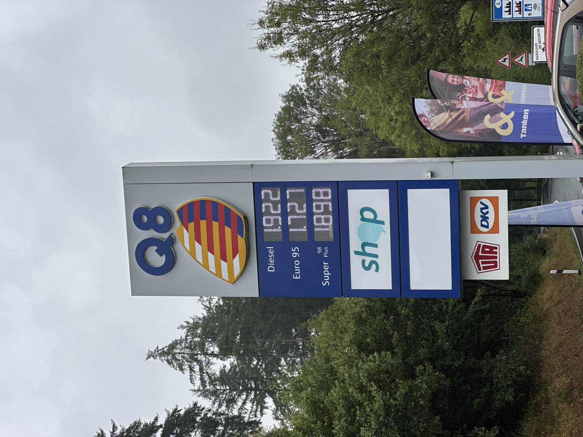 The image shows a Q8 gas station sign in Parc Hosingen, Luxembourg, on an overcast day. The sign displays fuel prices: Diesel at 1.922, Euro 95 at 1.727, and Super Plus 98 at 1.898. Below the prices, there is a blue panel with the word "shop" in white and teal. Further down are logos for UTA and DKV. Two large feather flags with images of people and the text "& Tanken & Shoppen" are visible to the right of the sign. Traffic signs, including a deer crossing warning, a speed limit sign, and a "Marquage de Sécurité" sign, are also present. Trees and foliage surround the gas station. A car is parked in front of the gas station.