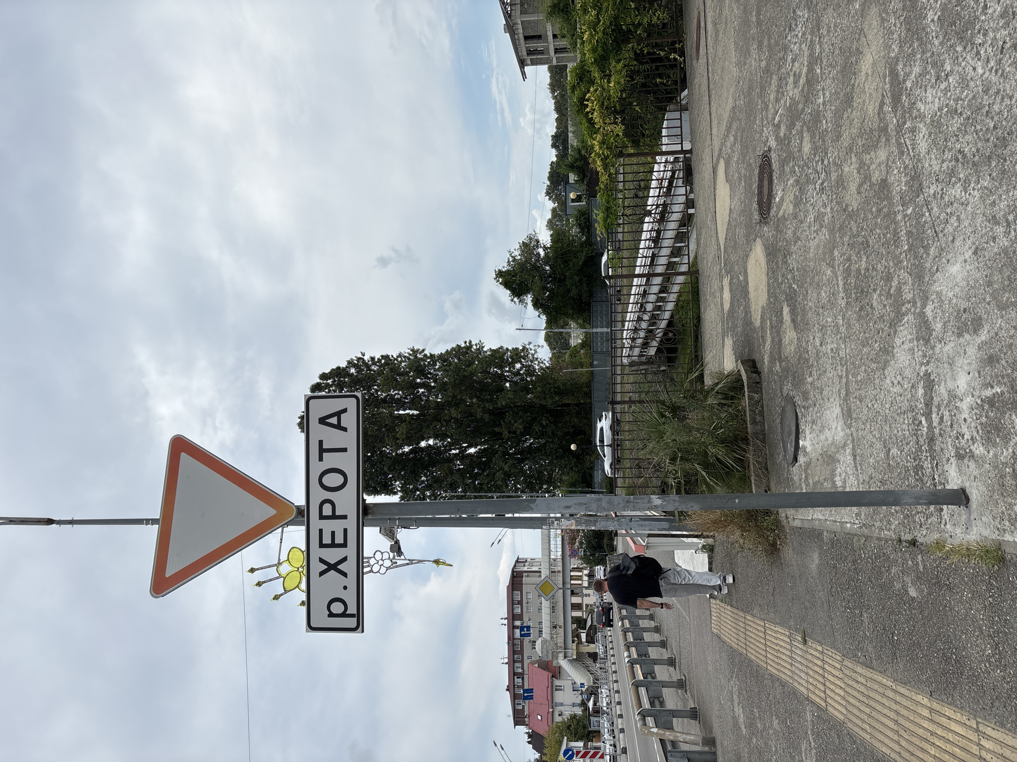 A yield sign and a street sign with the Cyrillic text "р. ХЕРОТА" are mounted on a pole. The street sign has a decorative yellow element resembling a butterfly or flower attached to it. A man walks away from the camera on a sidewalk, carrying a backpack. A yellow tactile paving strip runs alongside the sidewalk. In the background, there is a road with cars, buildings, trees, and a pedestrian bridge. The sky is cloudy. This scene is located in the city of Sochi, Russia, specifically on the road leading to Adler Airport.