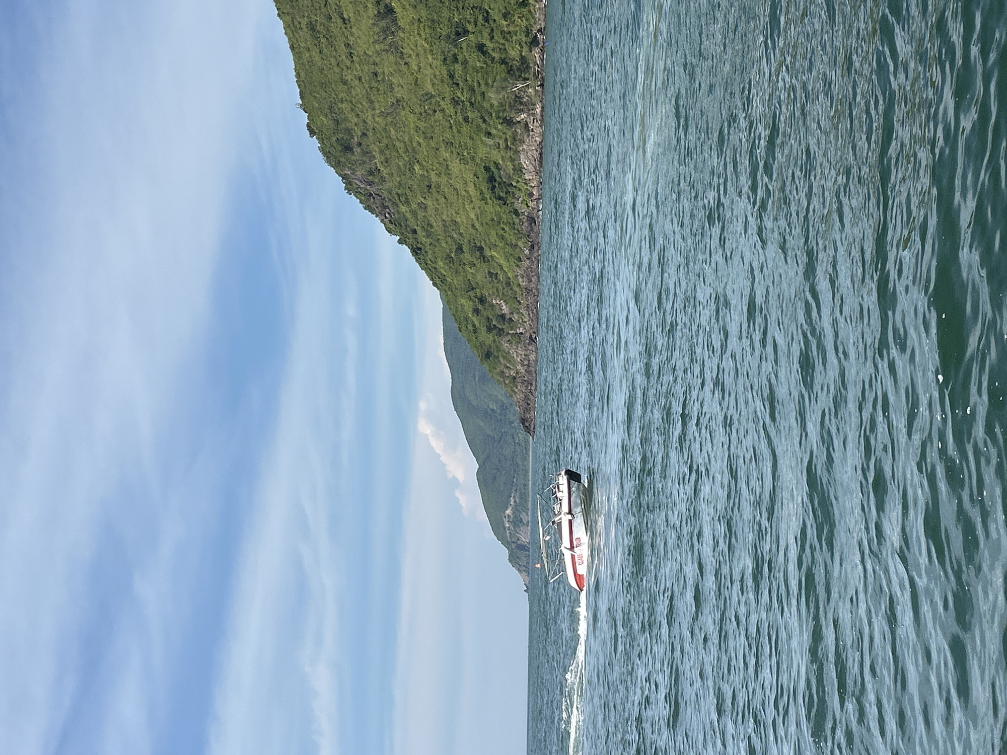 A white and red motorboat, identified by the inscription "CLEAR TOUR" on its side, cruises through the blue ocean waters off the coast of Nha Trang, Vietnam. The boat leaves a white wake behind it as it moves from left to right across the frame. In the background, lush green hills rise steeply from the rocky shoreline, creating a dramatic contrast with the expansive blue sky above, which is streaked with thin, wispy clouds. The scene captures a moment of leisure and travel along the Vietnamese coast.