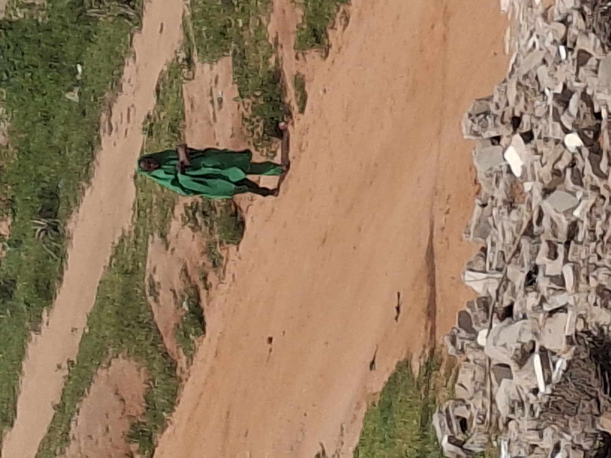 A person walks along a dirt road in a rural or semi-rural area. The person is wearing a green garment that covers most of their body and head, and they are carrying a dark object. The road is surrounded by dry grass and some vegetation. In the foreground, there is a pile of rubble, possibly from a collapsed structure. The image appears to be taken in Tumfure, Nigeria.