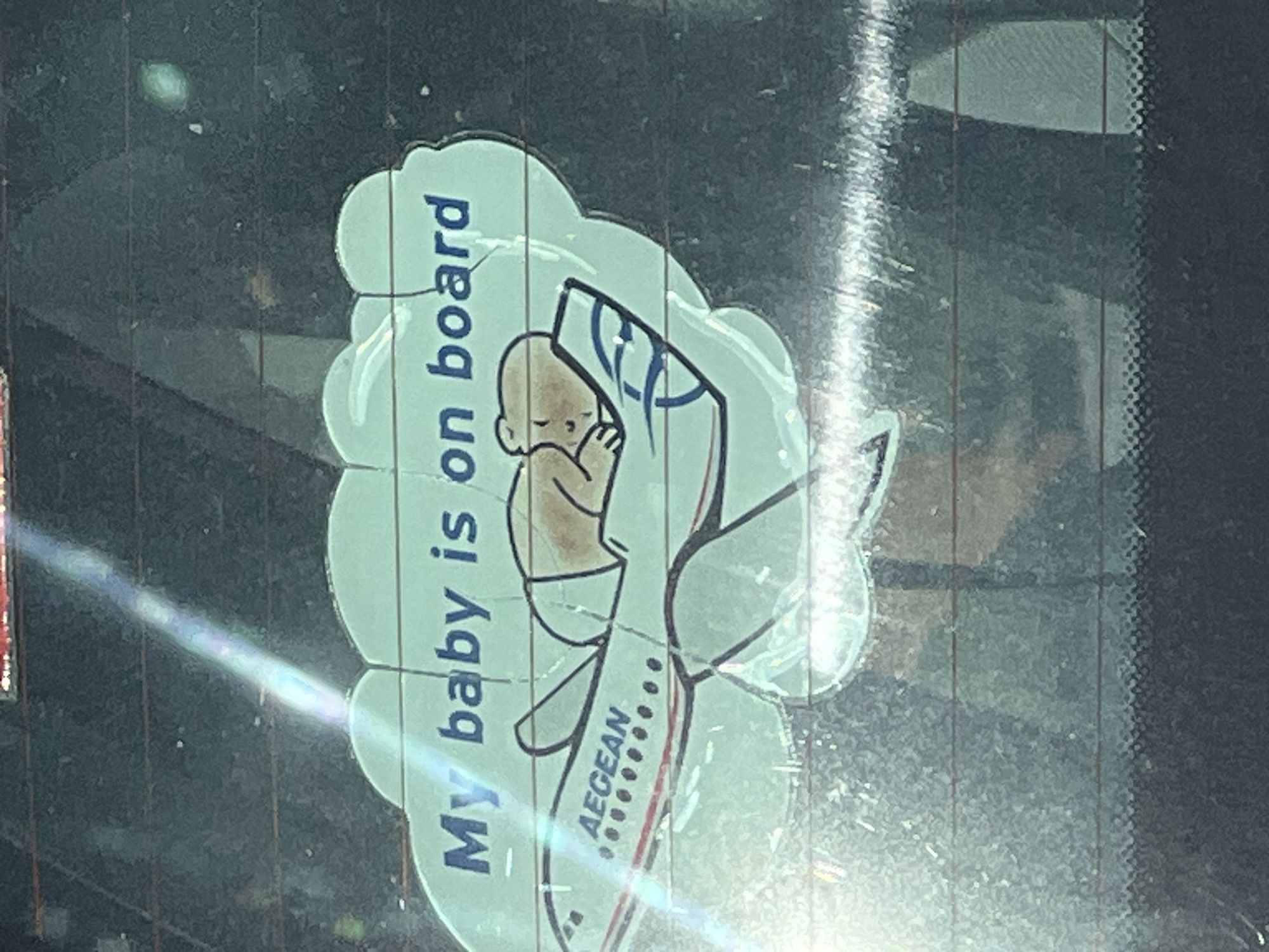 The image shows a "My baby is on board" sticker adhered to the inside of a car's rear window. The sticker features a cartoon baby nestled within the tail section of an Aegean Airlines plane. The sticker is light blue with black outlines and vertical text. The background is the dirty rear windshield of a car. The location is Plovdiv, Bulgaria.
