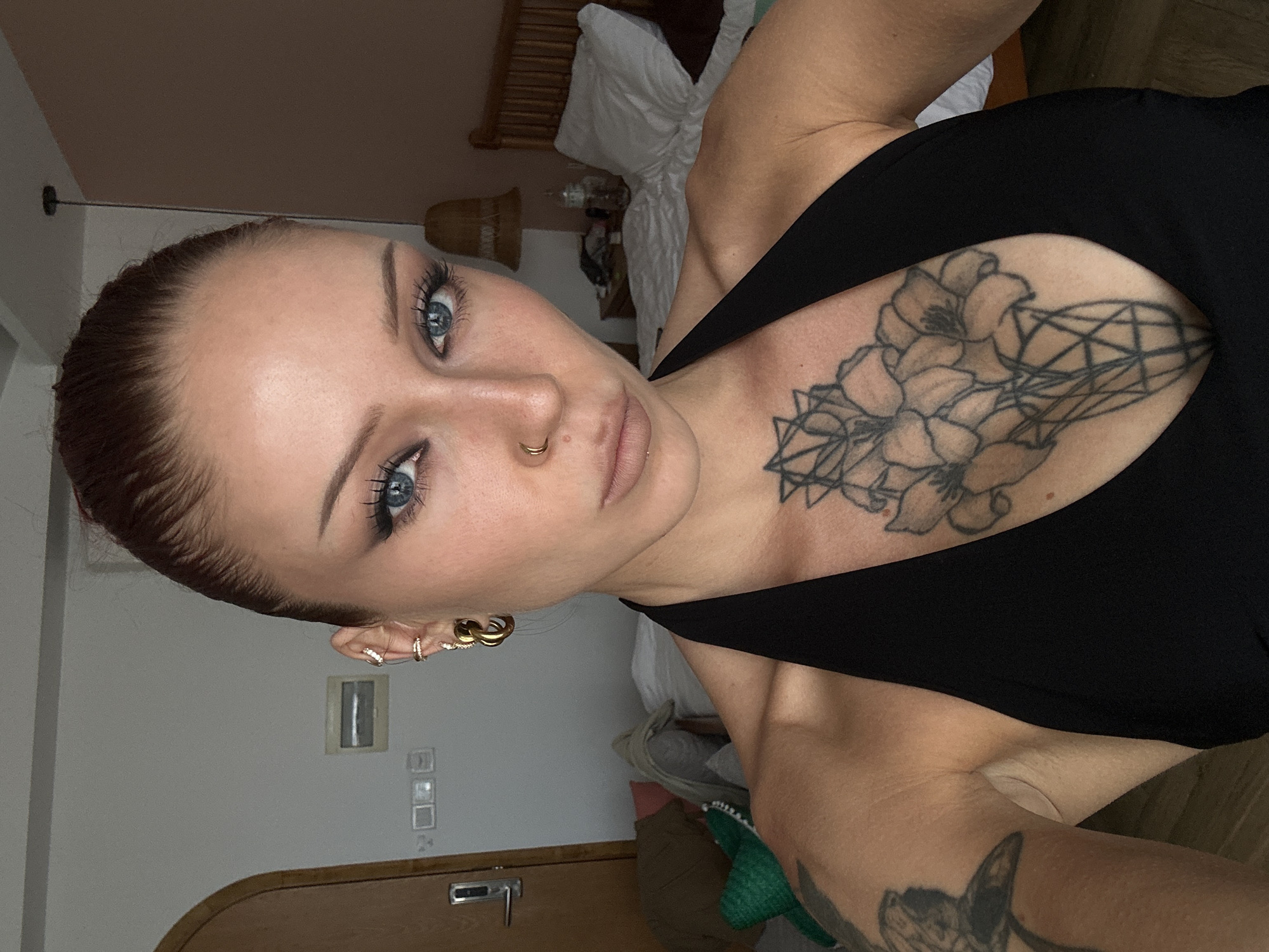 A woman with auburn hair pulled back and blue eyes is posing for a selfie indoors, likely in a hotel room in Da Nang, Vietnam. She is wearing a black halter top and has tattoos on her chest and left arm. She is also wearing hoop earrings and a nose ring. The background shows a bed, a wooden headboard, a bedside table with a lamp, and a door. The room appears to be somewhat messy, with clothes and bedding visible. The lighting is natural and comes from a window, casting shadows on the woman's face and body.