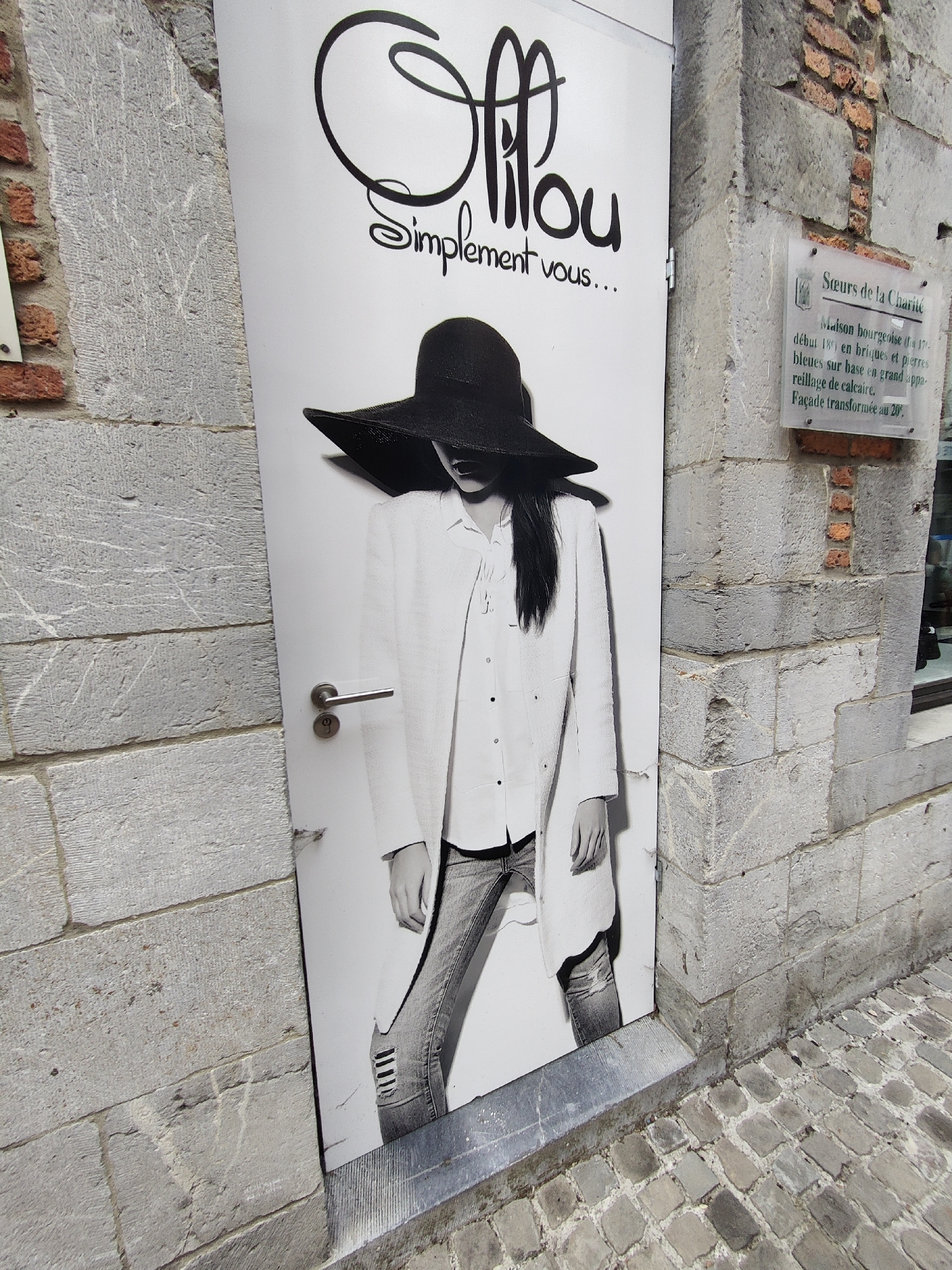 The image shows a black and white photograph of a woman wearing a wide-brimmed hat, a white coat, and ripped jeans. She is positioned on a door with a graphic printed on it, which appears to be an advertisement or a storefront. The door is set within a stone wall and is part of a building. The building's facade is made of rough-hewn stone blocks. To the right of the door, there is a glass plaque with text in French, indicating that the building is the "Sœurs de la Charité" (Sisters of Charity) and is a "Maison bourgeoise" (bourgeois house) dating from the 17th century, with a facade transformed in the 20th century. The text also mentions "briques et pierres bleues sur base en grand appareillage de calcaire" (bricks and blue stones on a base of large limestone masonry). The ground in front of the building is paved with cobblestones. The location is Marche-en-Famenne, Belgium.