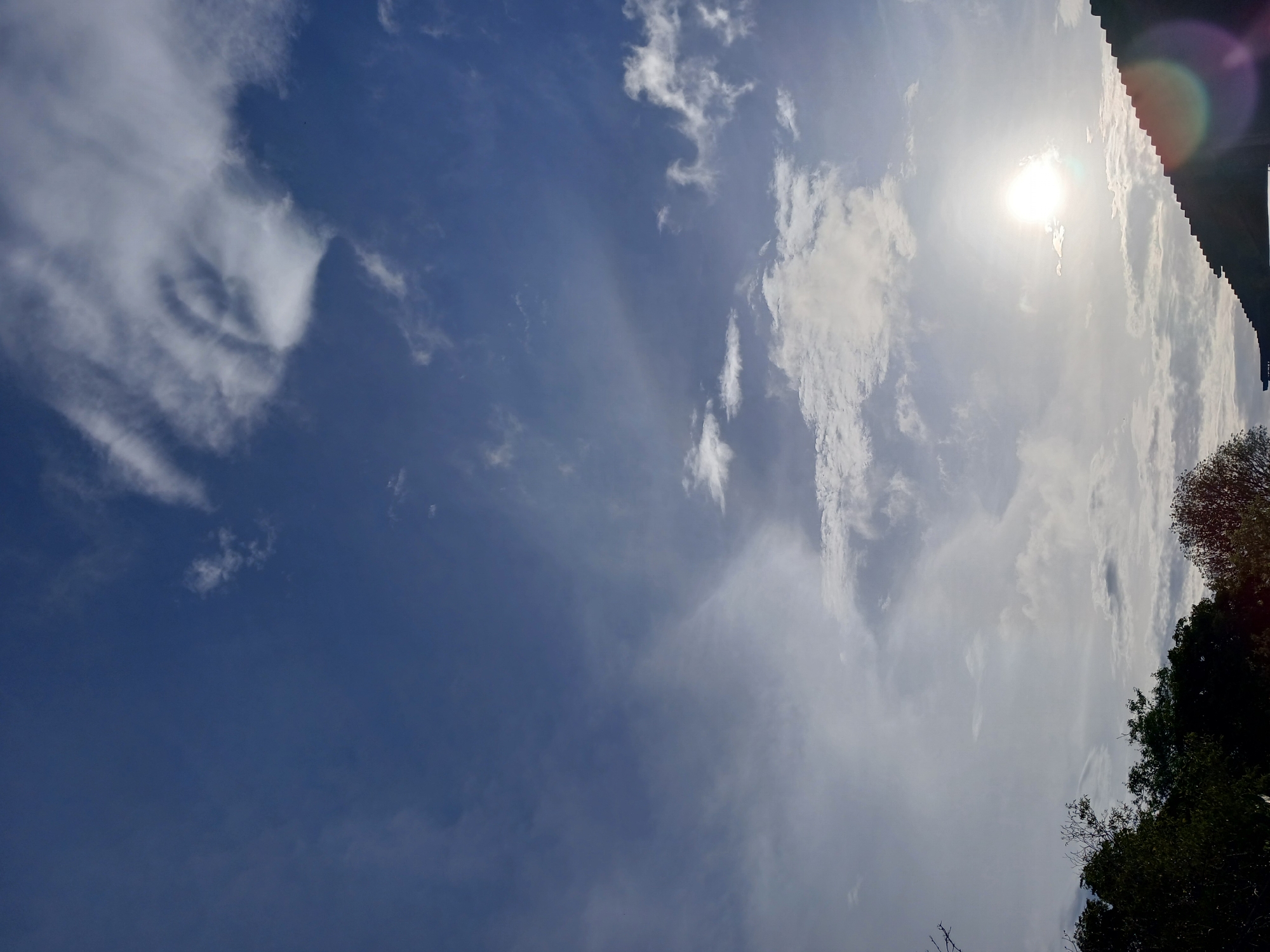 The image depicts a sky filled with clouds over Altagracia, Venezuela. The sun is visible as a bright orb in the lower right quadrant of the image, partially obscured by clouds and casting lens flare. Silhouetted against the bright sky in the lower portion of the image are the dark shapes of trees and the edge of a roofline. The clouds are varied in texture and density, with some appearing wispy and others more solid. The overall impression is that of an outdoor scene with natural lighting conditions typical of a sunny day.