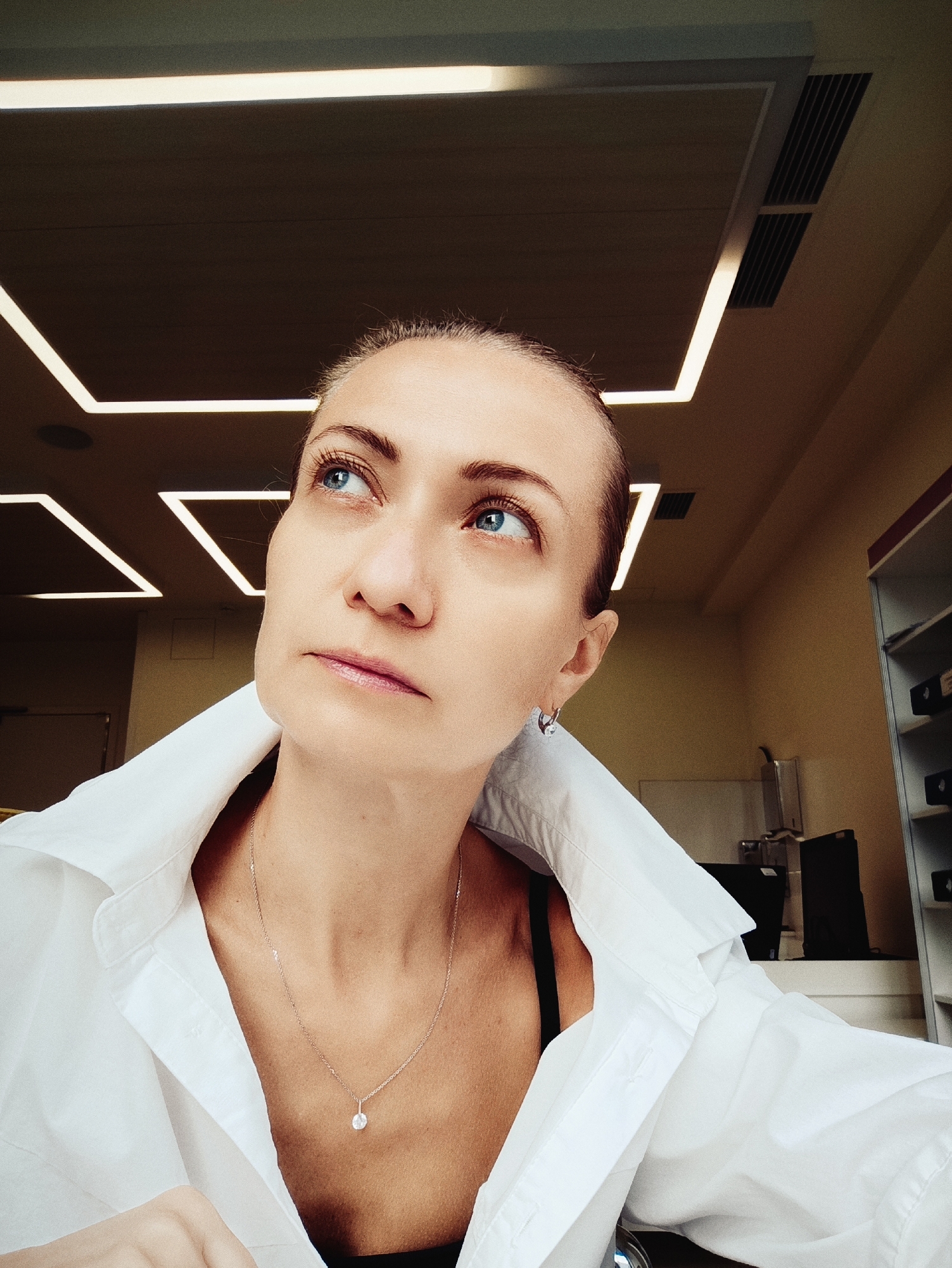 A close-up, high-angle shot focuses on a woman looking upwards and to her left. She has fair skin, blue eyes, and her hair is pulled back. She is wearing a white button-down shirt with the collar open, revealing a glimpse of a black bra and a delicate silver necklace with a pendant. The background suggests an indoor setting with modern, geometric lighting fixtures on a dark ceiling and beige walls. The lighting is warm and somewhat dim. No specific location in Moscow, Russia is identifiable from the image.
