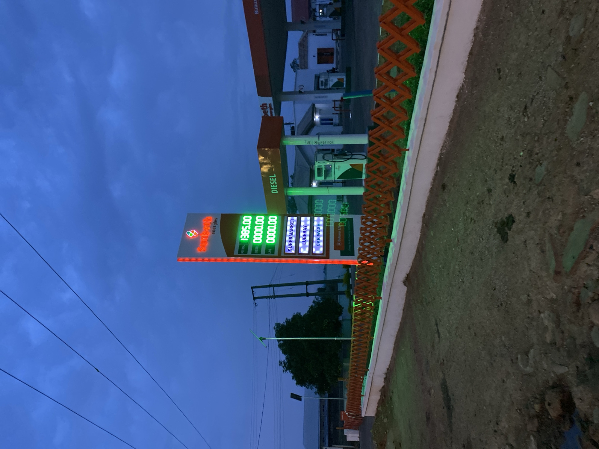 The image depicts a gas station in Kuru Dungus, Nigeria at dusk. The gas station sign advertises "Sunbeth energies" and displays fuel prices: "PETROL 1385.00", "DIESEL 0000.00", and "LPG 0000.00". The sign also lists "SUPERMARKET", "LUBEBAY", and "CAR WASH". Gas pumps are visible under the station's canopy. The sky is a deep blue. Power lines run across the lower part of the sky. A low wall with an orange fence runs along the edge of the property. The ground is a mixture of dirt and gravel.